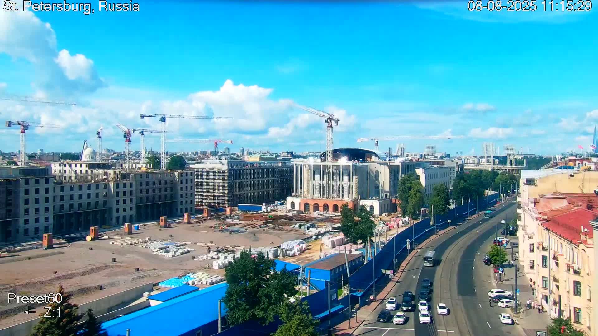 A partly cloudy sunny day reveals an expansive construction site with numerous cranes and buildings under development, adjacent to a busy city street.