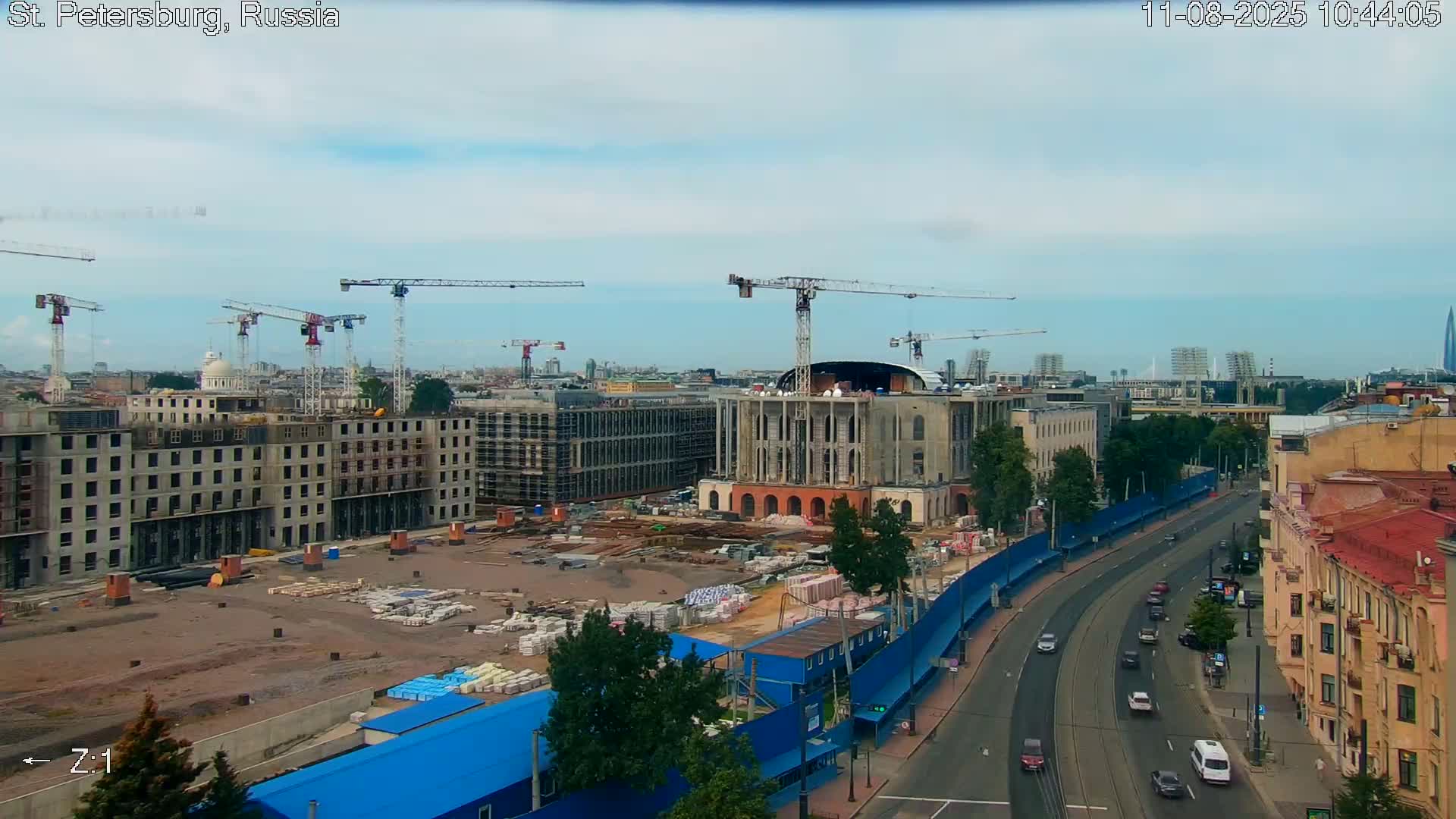 A large-scale construction site with multiple cranes and buildings under construction is visible, alongside a roadway with cars, under a partly cloudy sky.