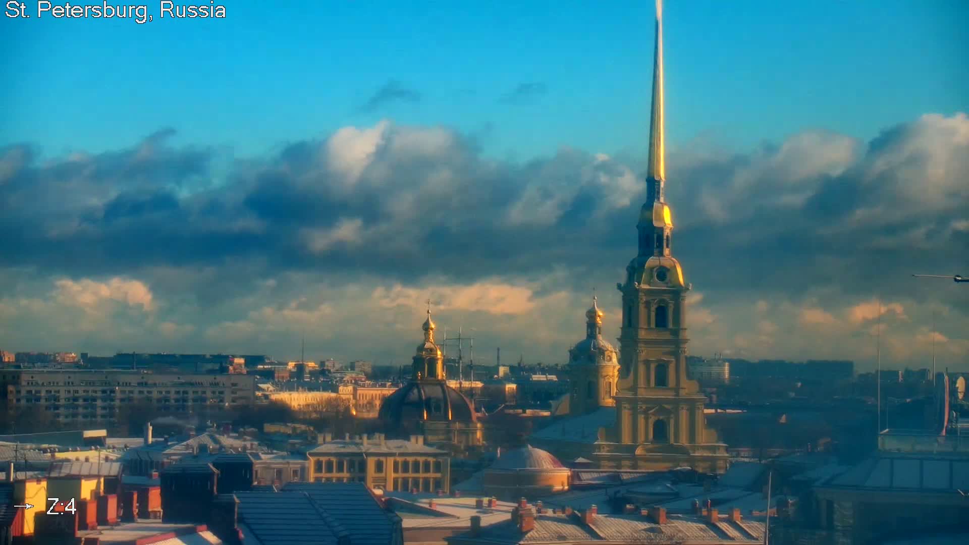 A city skyline featuring numerous golden spires and domes, most notably a tall, slender gilded spire, extends under a partly cloudy blue sky, with many rooftops showing a light covering of snow, indicative of cold weather.