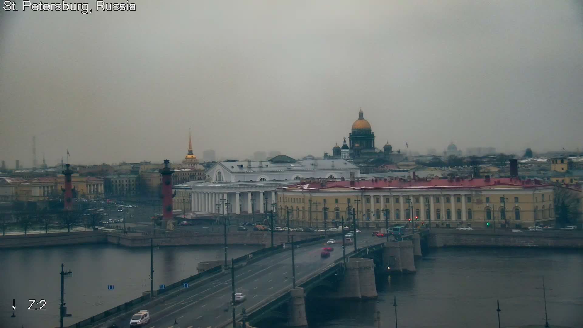A city skyline featuring numerous golden spires and domes, most notably a tall, slender gilded spire, extends under a partly cloudy blue sky, with many rooftops showing a light covering of snow, indicative of cold weather.