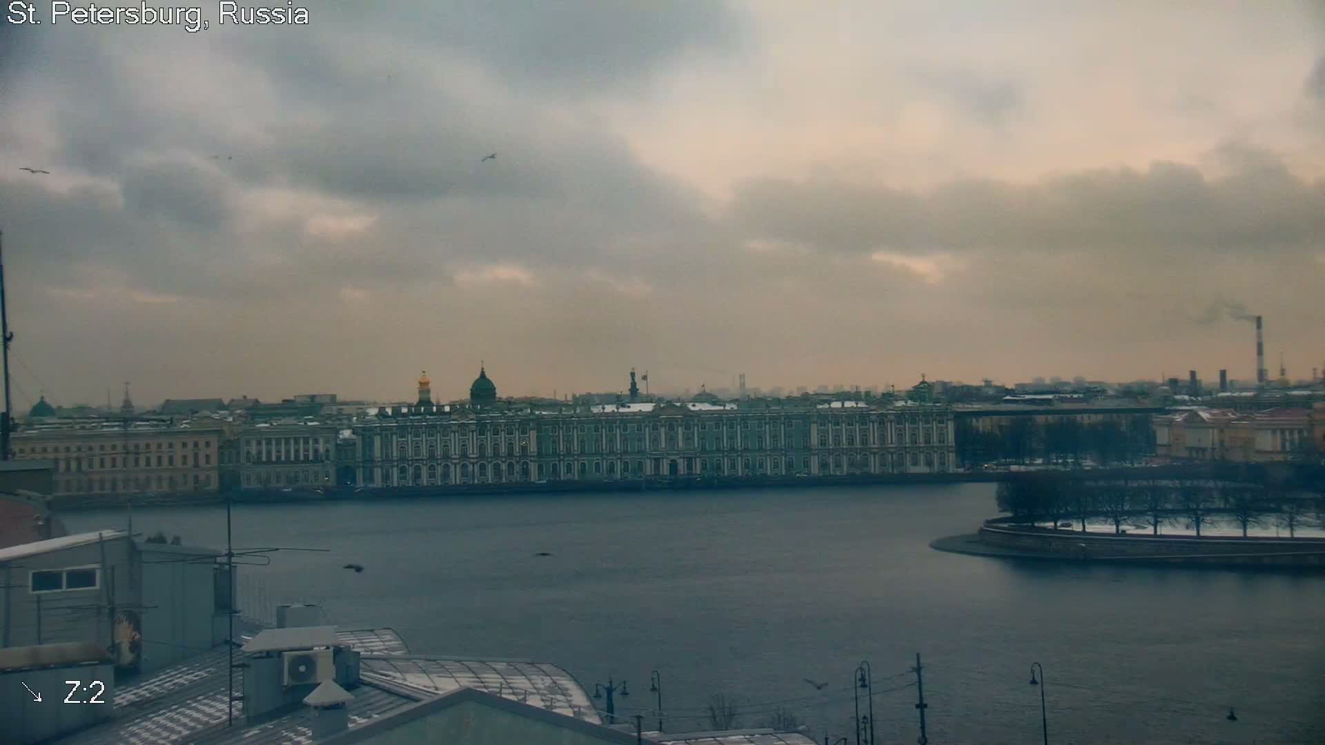 A wide river flows past a grand city skyline featuring ornate buildings and a tree-lined island, viewed from above snowy rooftops on an overcast and grey winter day.