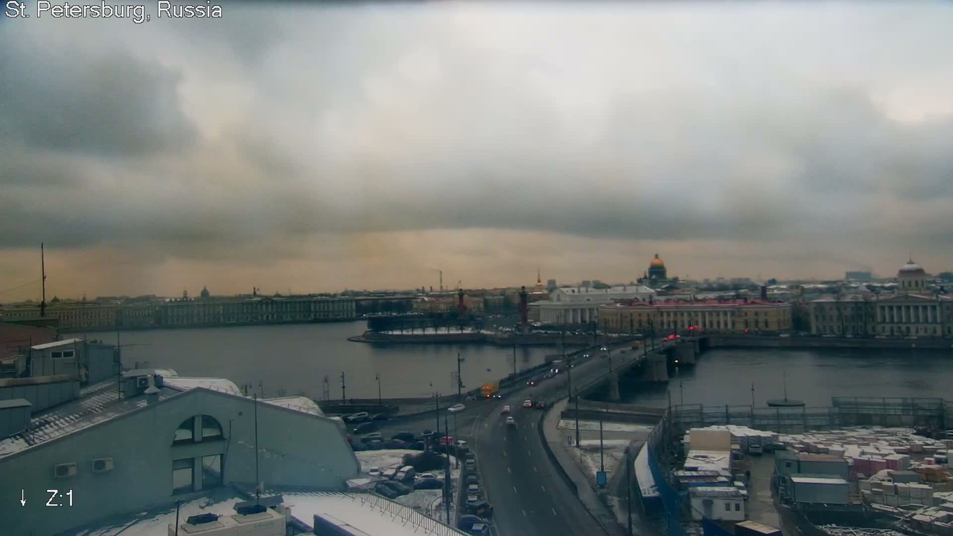 A wide river flows past a grand city skyline featuring ornate buildings and a tree-lined island, viewed from above snowy rooftops on an overcast and grey winter day.