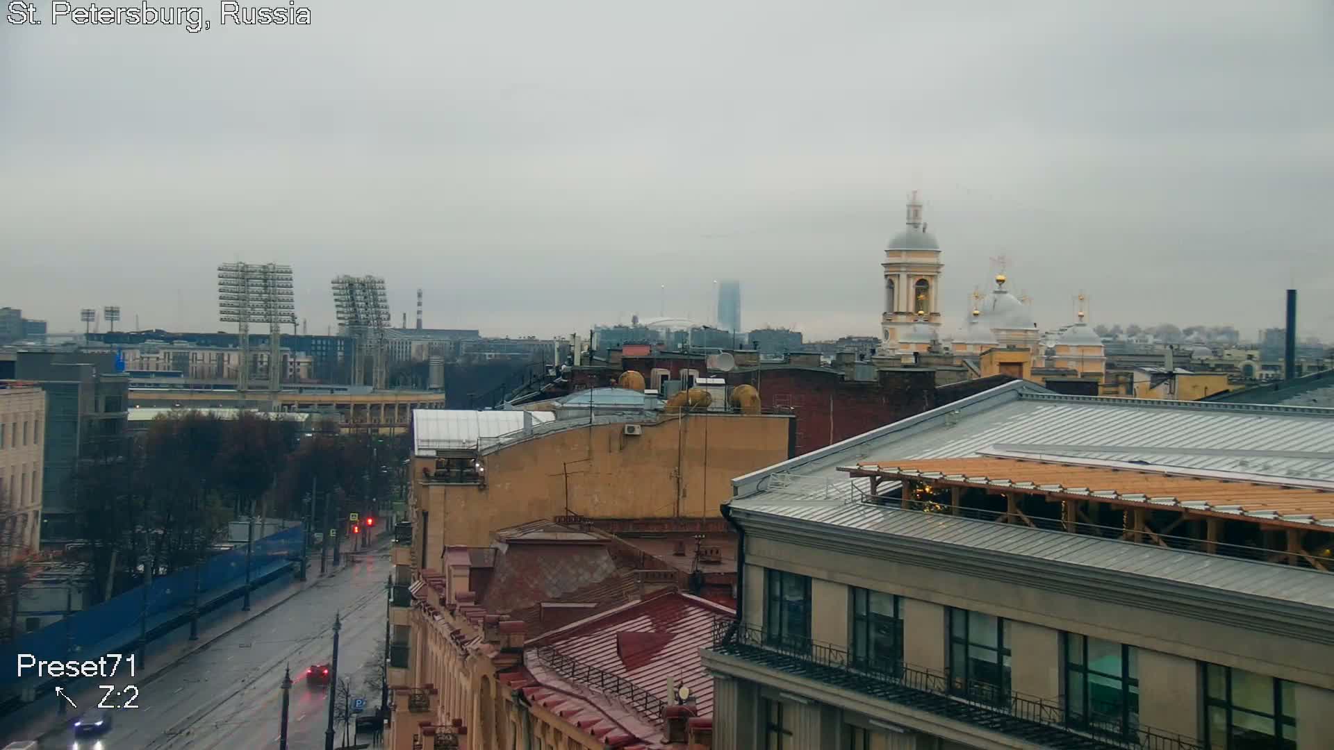 A wide urban view on an overcast and wet day reveals a street with cars, varied buildings with rooftops, a stadium with floodlights, distant domed churches, and a tall skyscraper against a grey sky.