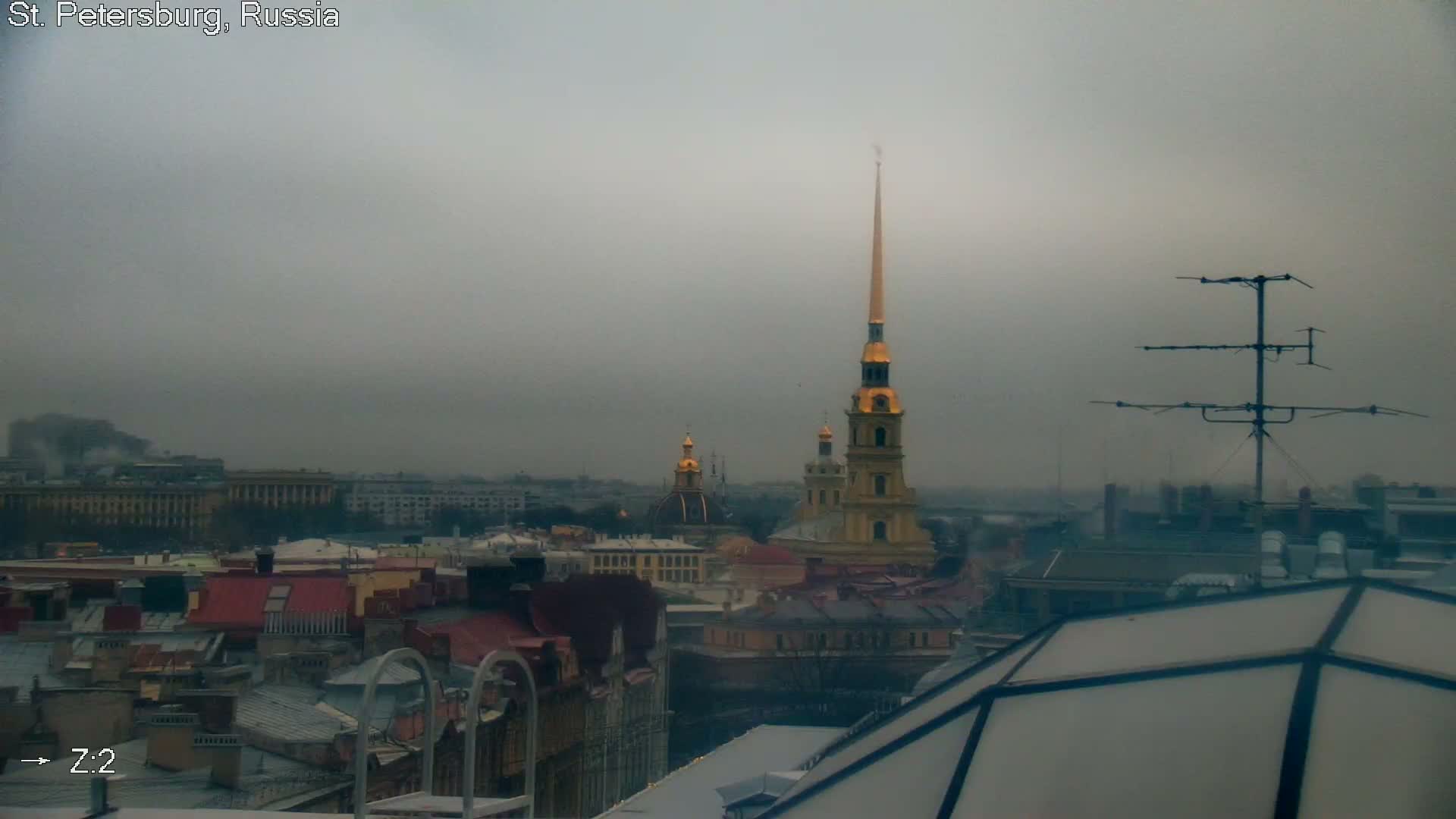 An overcast and hazy view of a city skyline features rooftops in the foreground, with a prominent golden-spired cathedral rising above other buildings in the midground, all under a dull grey sky.