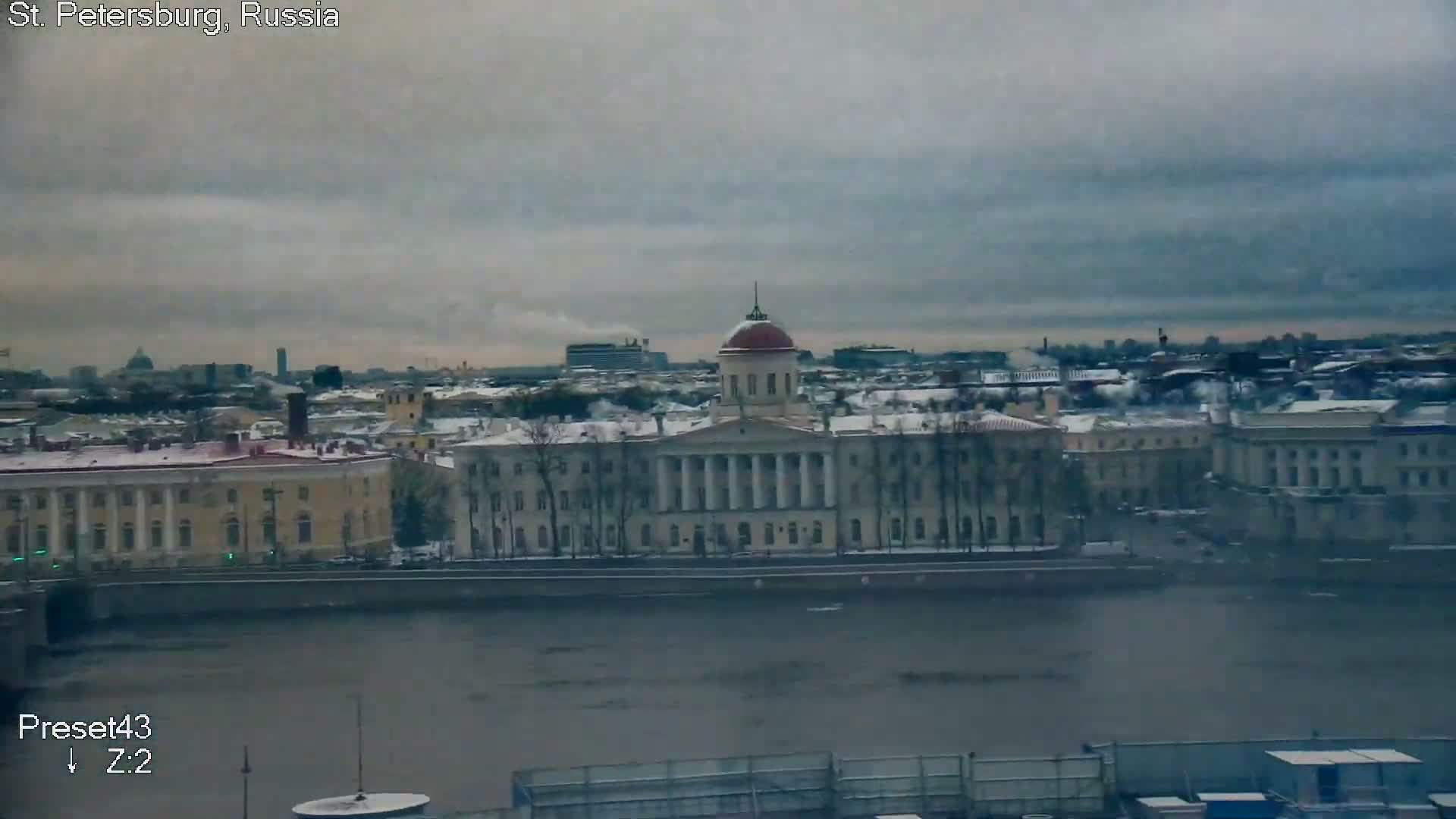 A panoramic view captures a cityscape with snow-dusted rooftops and classical buildings, including one with a prominent red dome, lining a wide river under a heavy, overcast grey sky on a cold winter day.