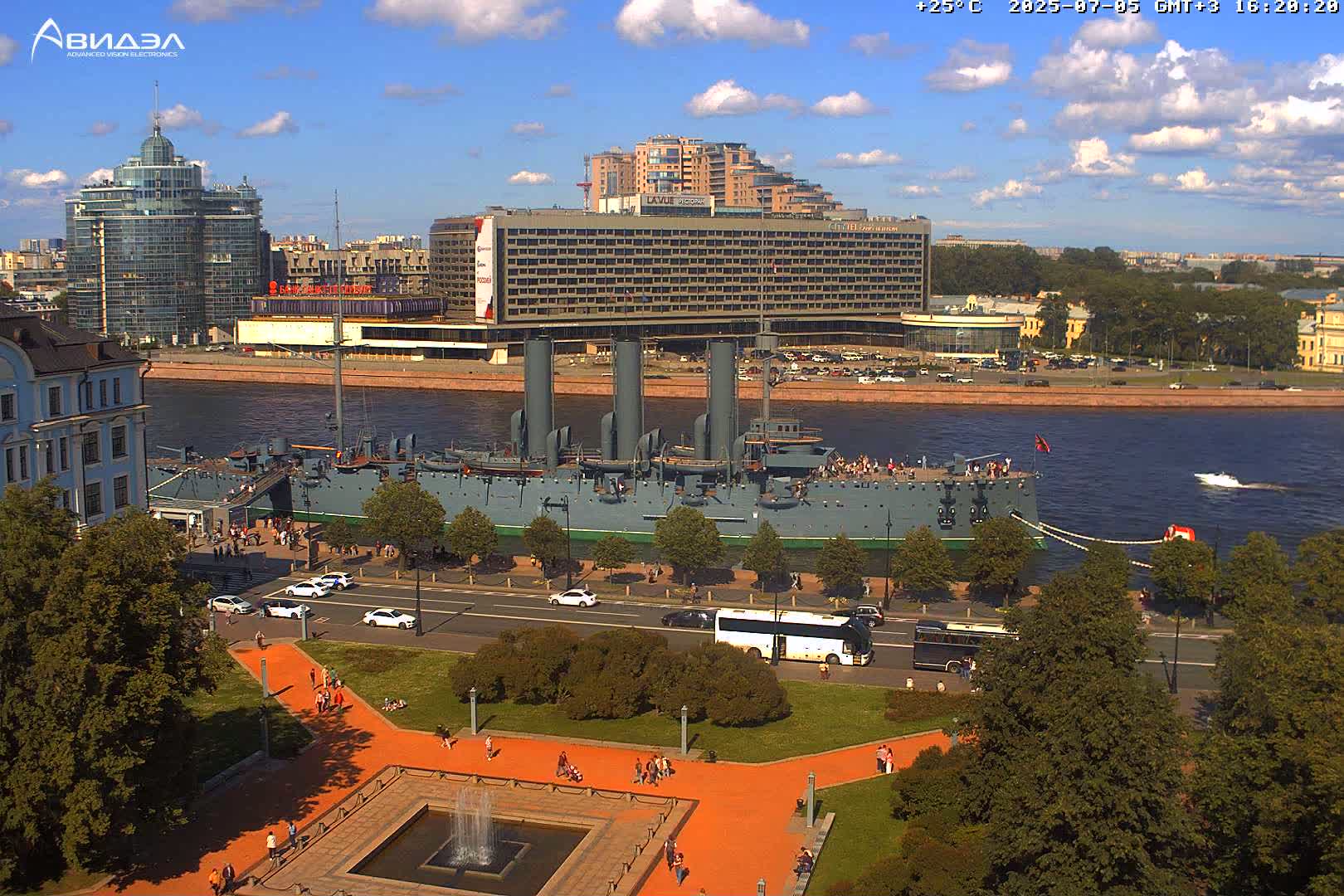 A large battleship is docked along a city riverfront under a mostly sunny sky, with people walking along a paved path next to a park and several buildings in the background.