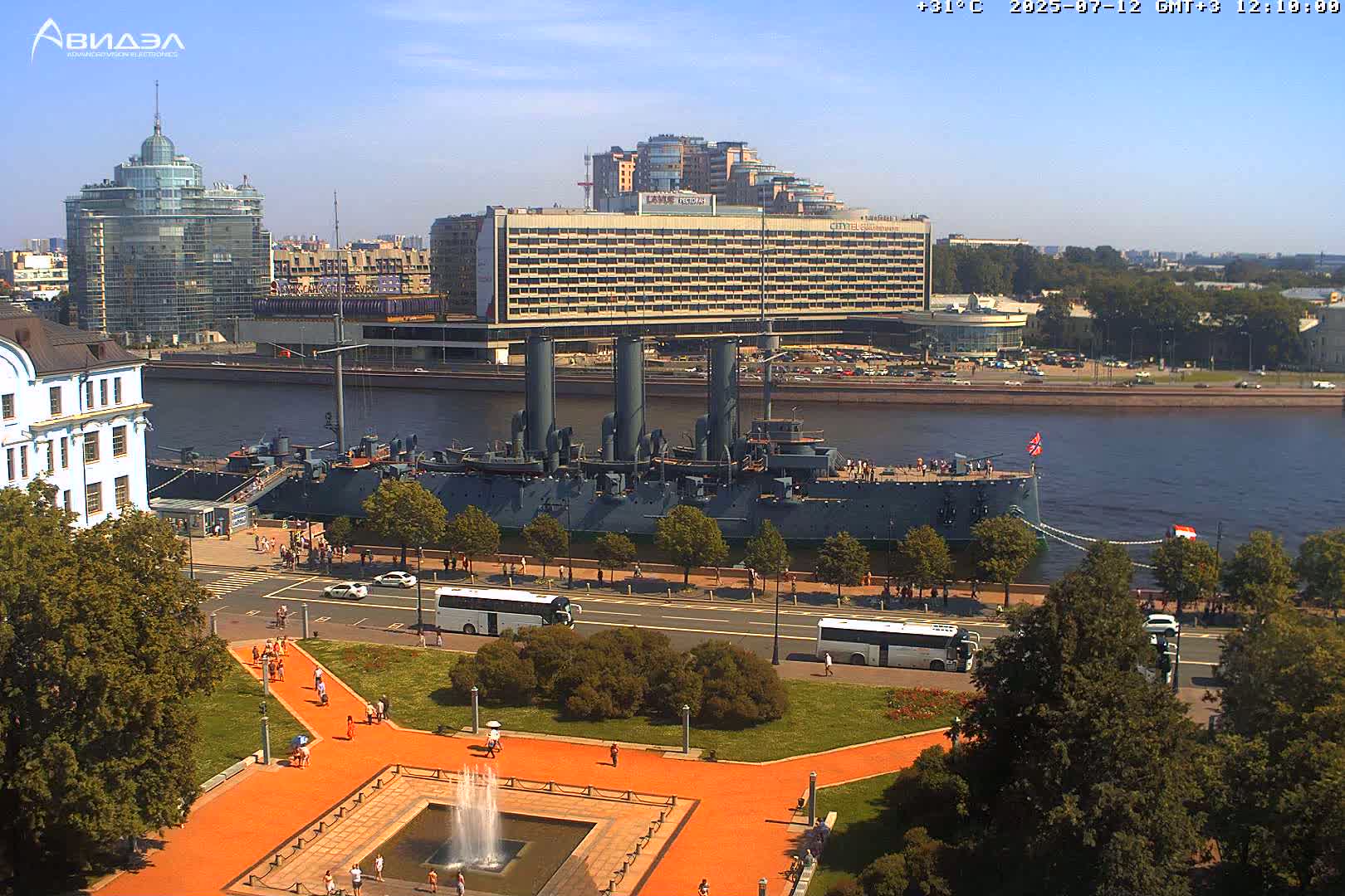 A large battleship is docked along a city riverfront under a mostly sunny sky, with people walking along a paved path next to a park and several buildings in the background.