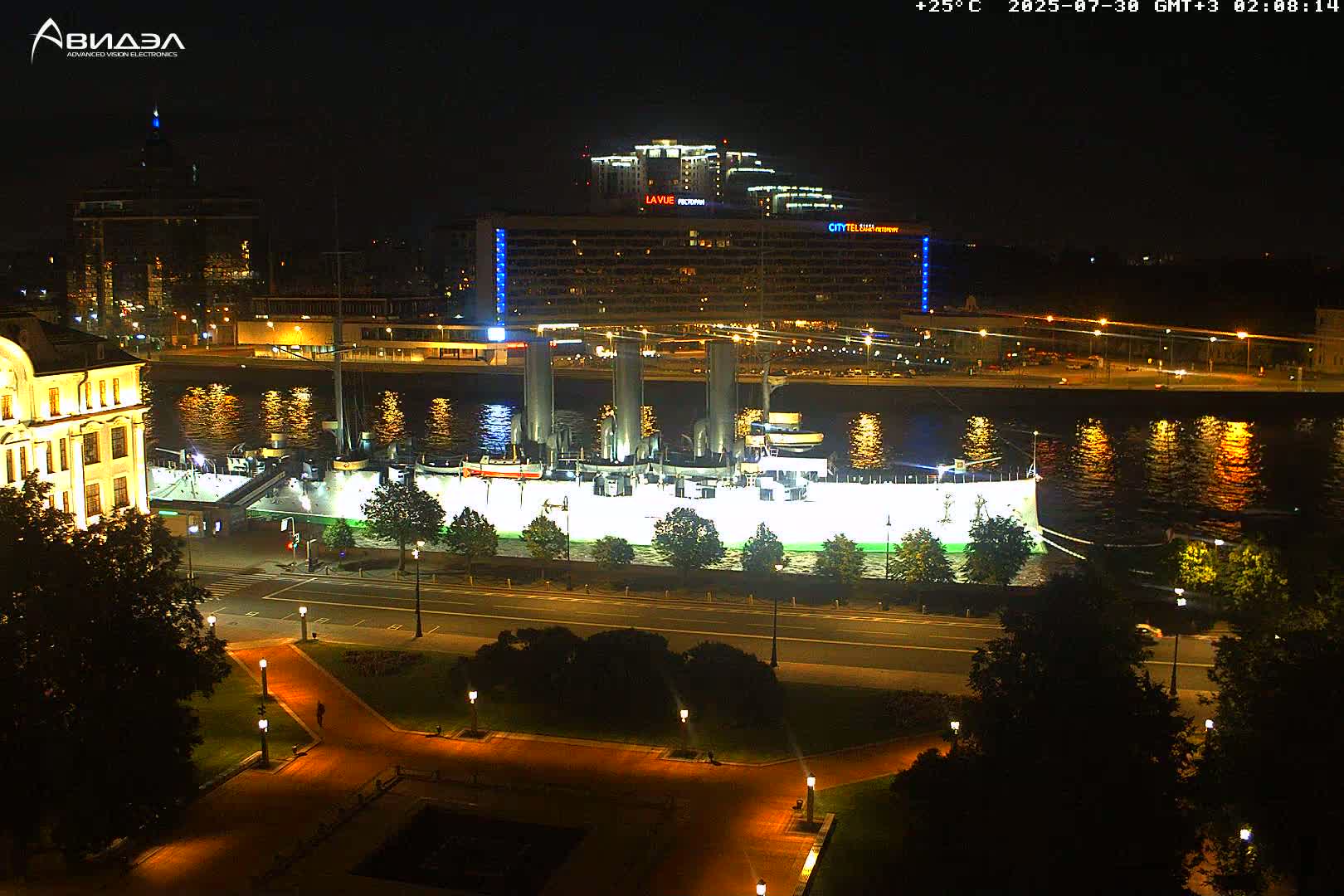 A large, dark-grey battleship is docked along a river under a clear, sunny sky; a park with a fountain is in the foreground, and several buses are parked along the riverbank.