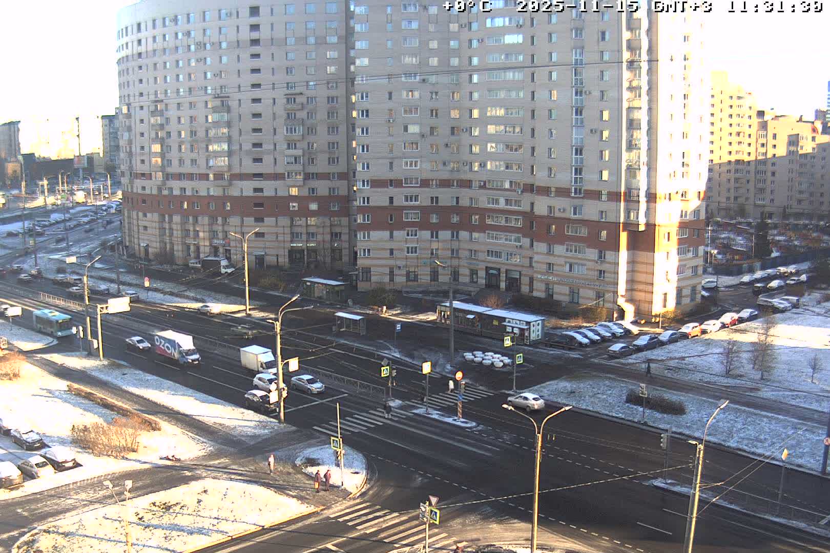 An urban street scene on a bright, cold day shows a thin layer of snow covering the ground and parked cars, with multiple lanes of traffic and large apartment buildings in the background.