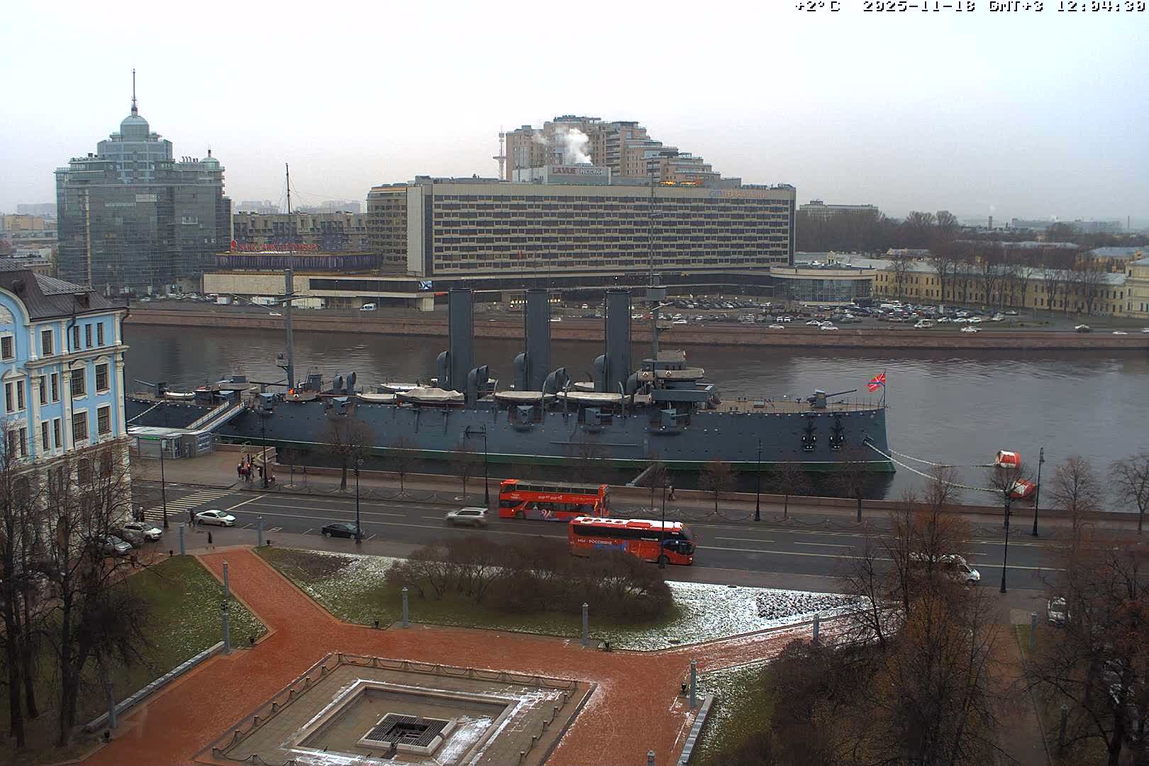 An urban street scene on a bright, cold day shows a thin layer of snow covering the ground and parked cars, with multiple lanes of traffic and large apartment buildings in the background.