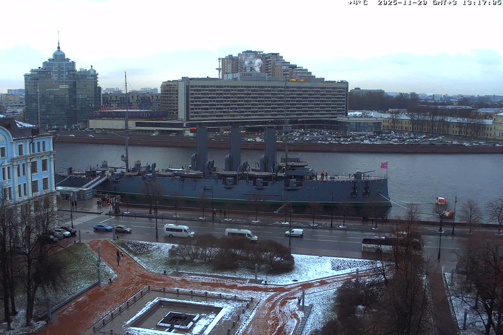 An overcast aerial view shows a historic warship docked on a wide river in an urban setting, with wet roads and patchy snow covering the ground and bare trees, indicating cold weather.