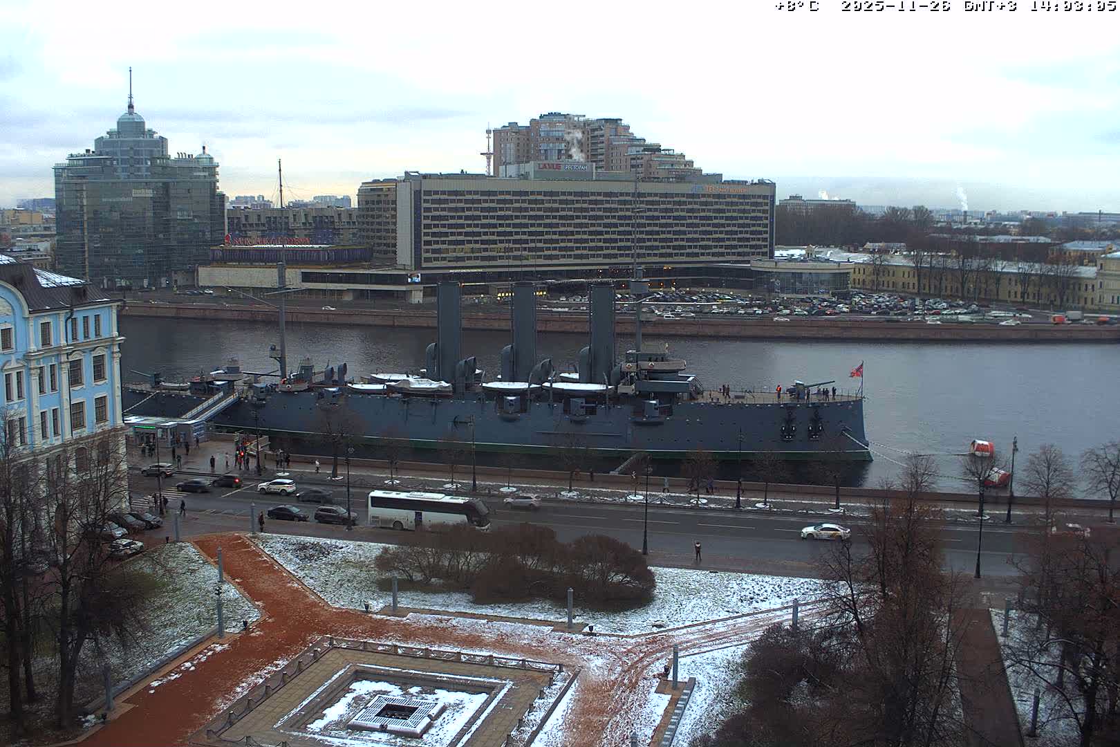 An overcast aerial view shows a historic warship docked on a wide river in an urban setting, with wet roads and patchy snow covering the ground and bare trees, indicating cold weather.