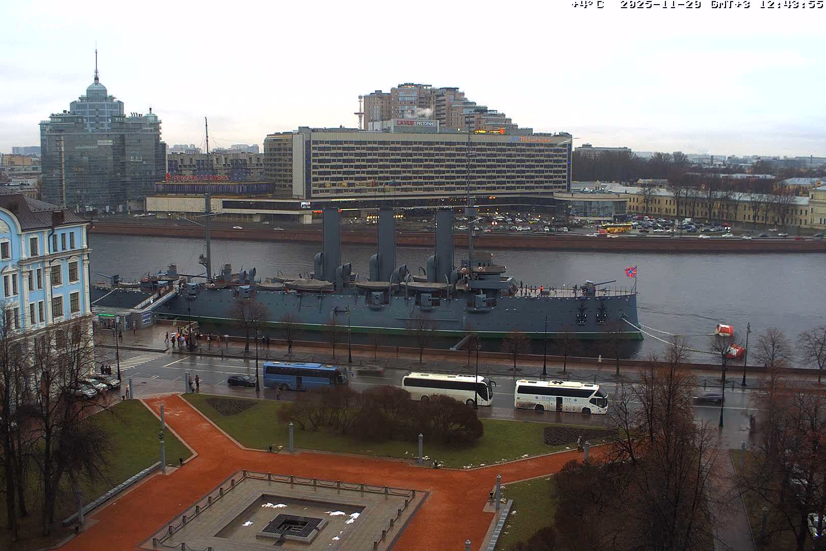 Under an overcast sky on a cool day, a historic grey warship is prominently moored on a wide river, bordered by a bustling urban landscape featuring modern and classic buildings, wet streets with buses and cars, and a park with bare trees.