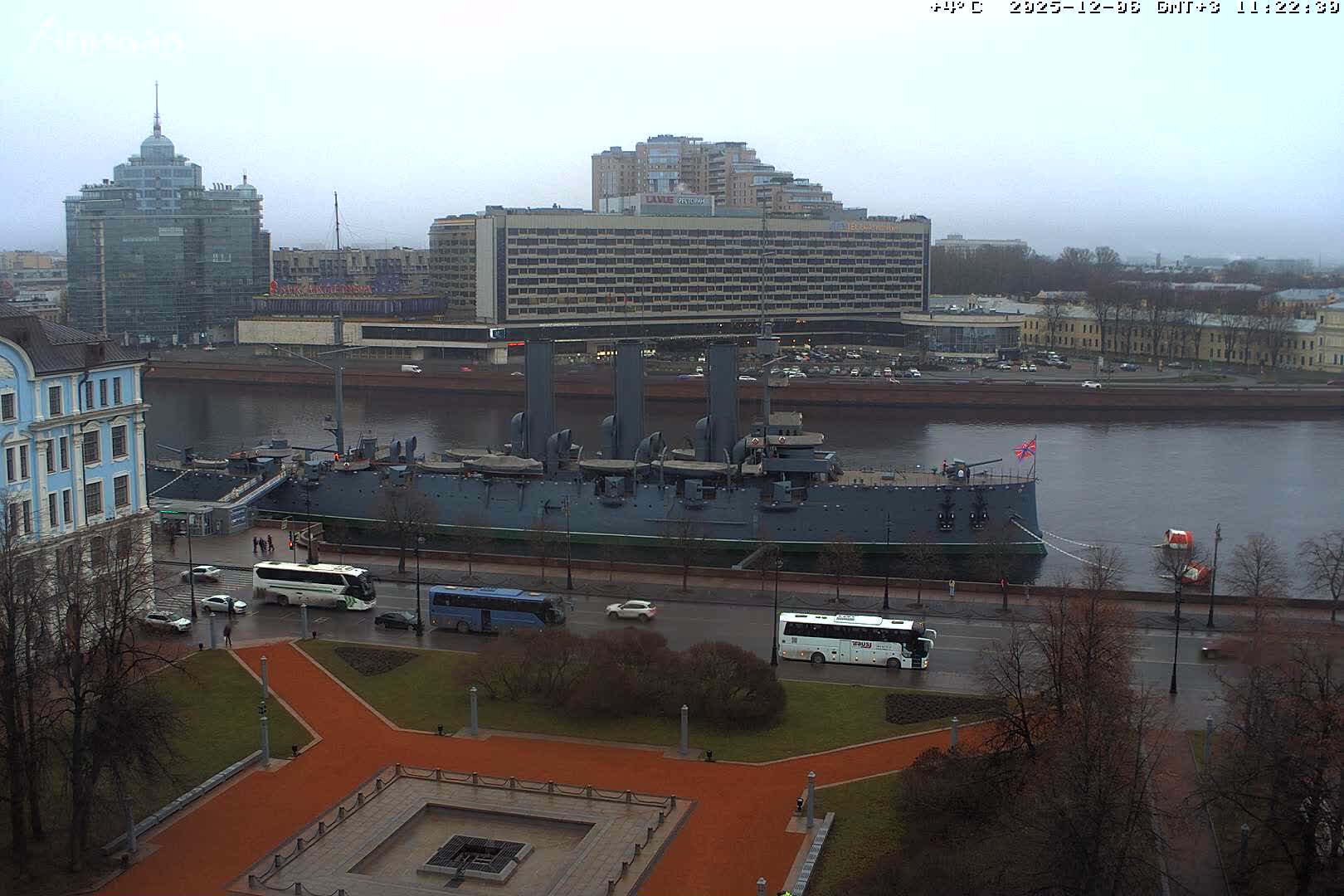 A large grey battleship is docked on a wide river in an urban setting under an overcast and damp sky, with a park and wet roads featuring buses and cars in the foreground, and numerous buildings in the background.
