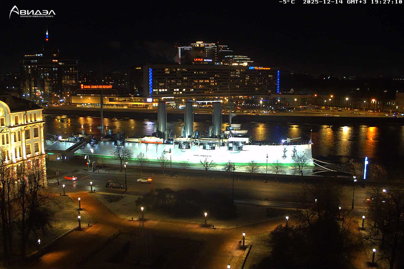 A large grey battleship is docked on a wide river in an urban setting under an overcast and damp sky, with a park and wet roads featuring buses and cars in the foreground, and numerous buildings in the background.