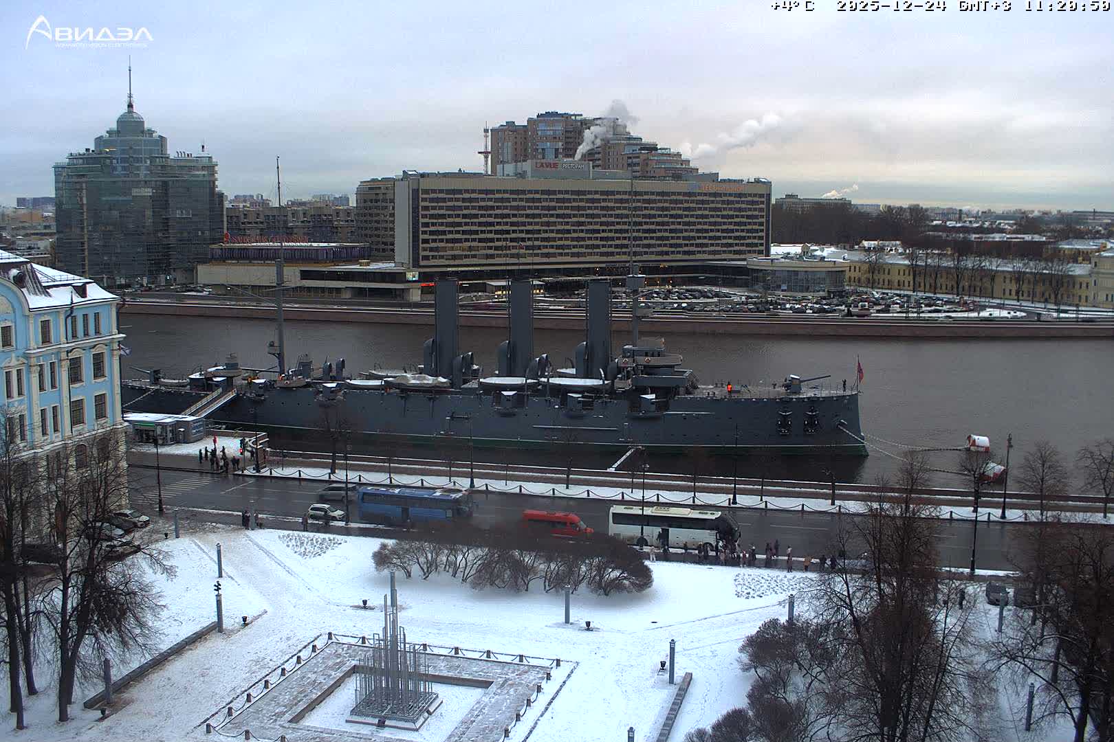 A snow-covered city embankment with a battleship docked on a wide river, surrounded by bare trees, traffic, and urban buildings under an overcast sky.
