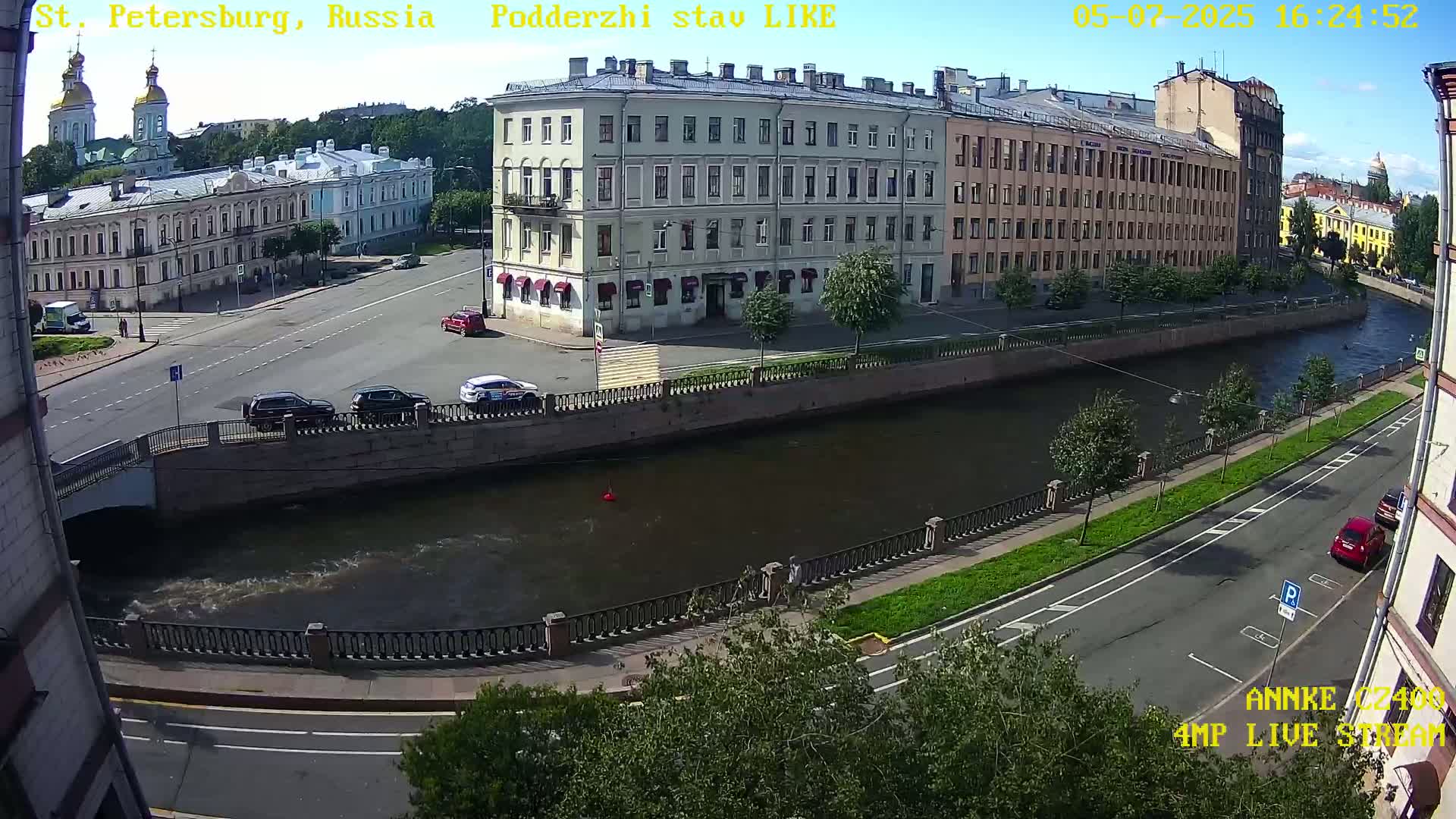 A sunny day in a city shows several buildings along a canal with cars parked and driving on streets nearby.