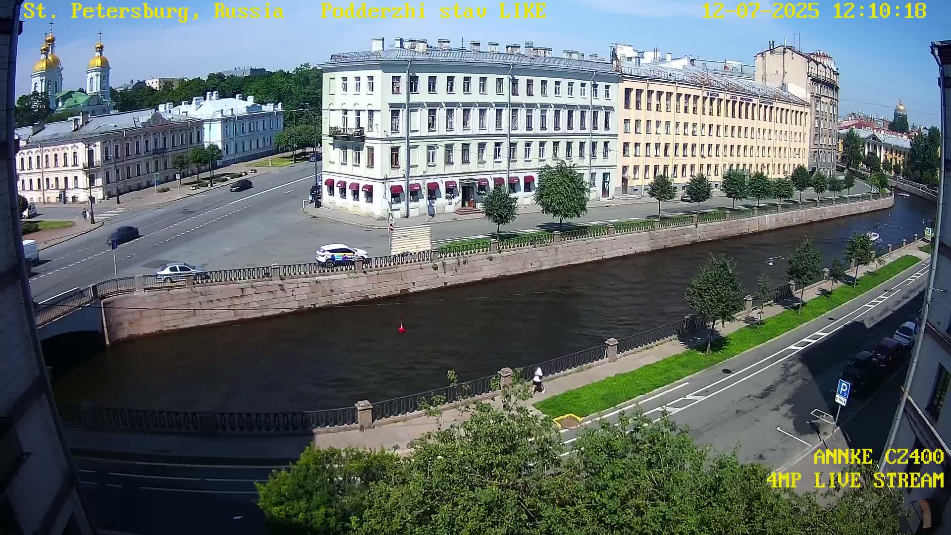A sunny day in a city shows several buildings along a canal with cars parked and driving on streets nearby.