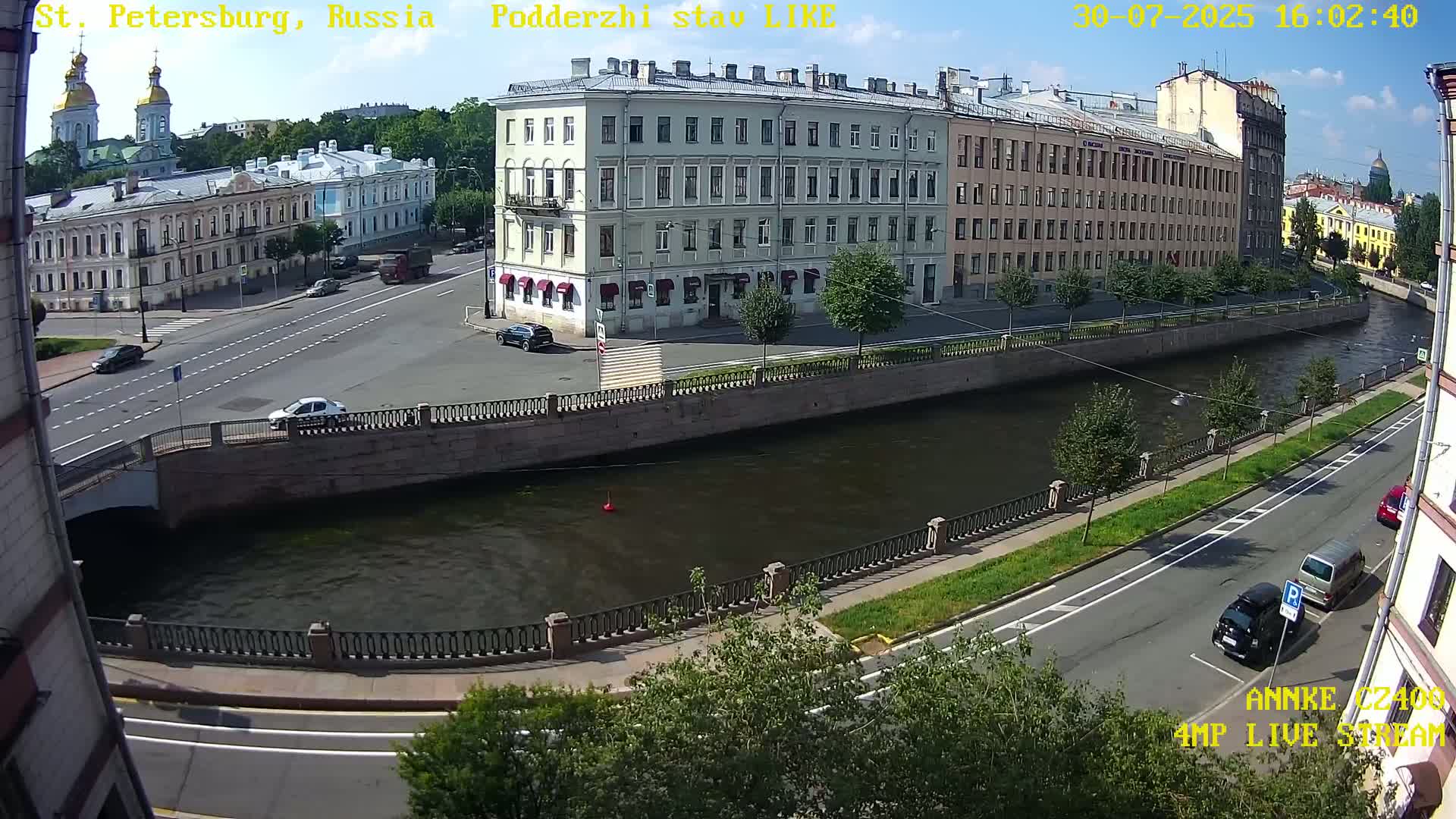 A sunny day overlooking a canal in a city with several multi-story buildings, cars on roads and a bridge.