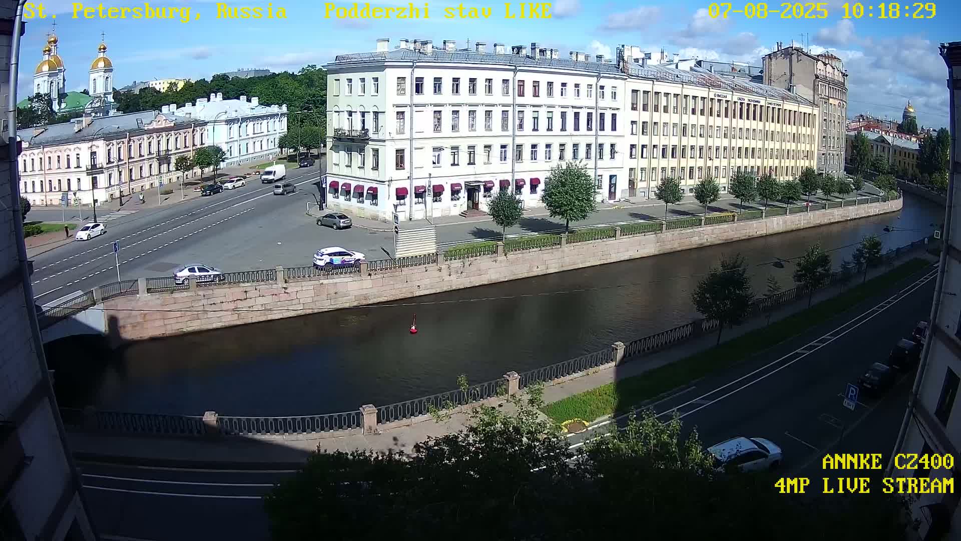 A sunny day in a city shows a canal with several buildings, cars, and a bridge, viewed from a high vantage point.