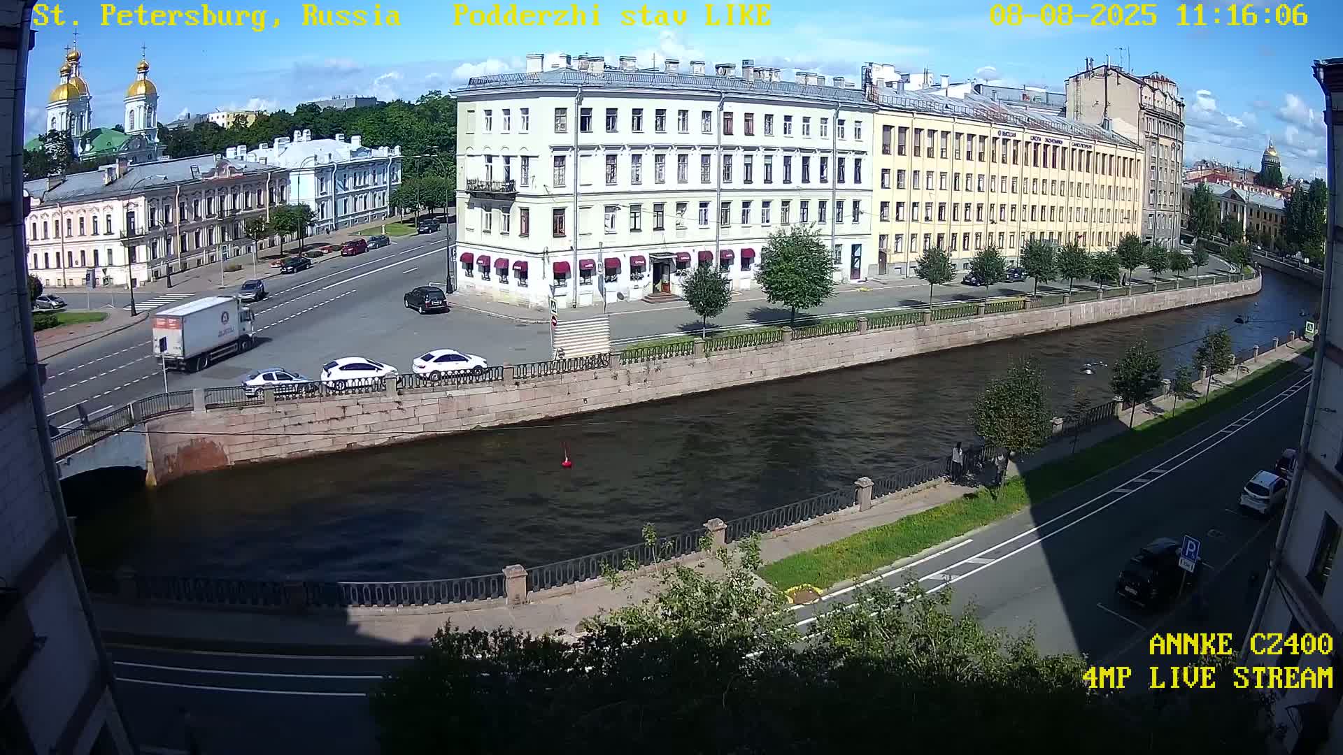 A sunny day shows a waterway with buildings lining the banks, cars on roads nearby, and a bridge crossing over the water.