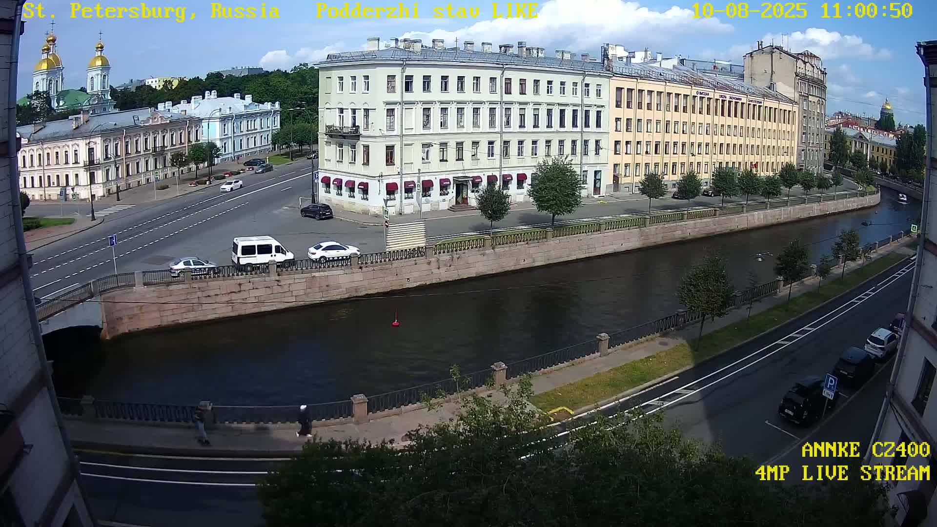 A sunny day in a city shows a canal with buildings and vehicles lining its banks, and a bridge crossing it.