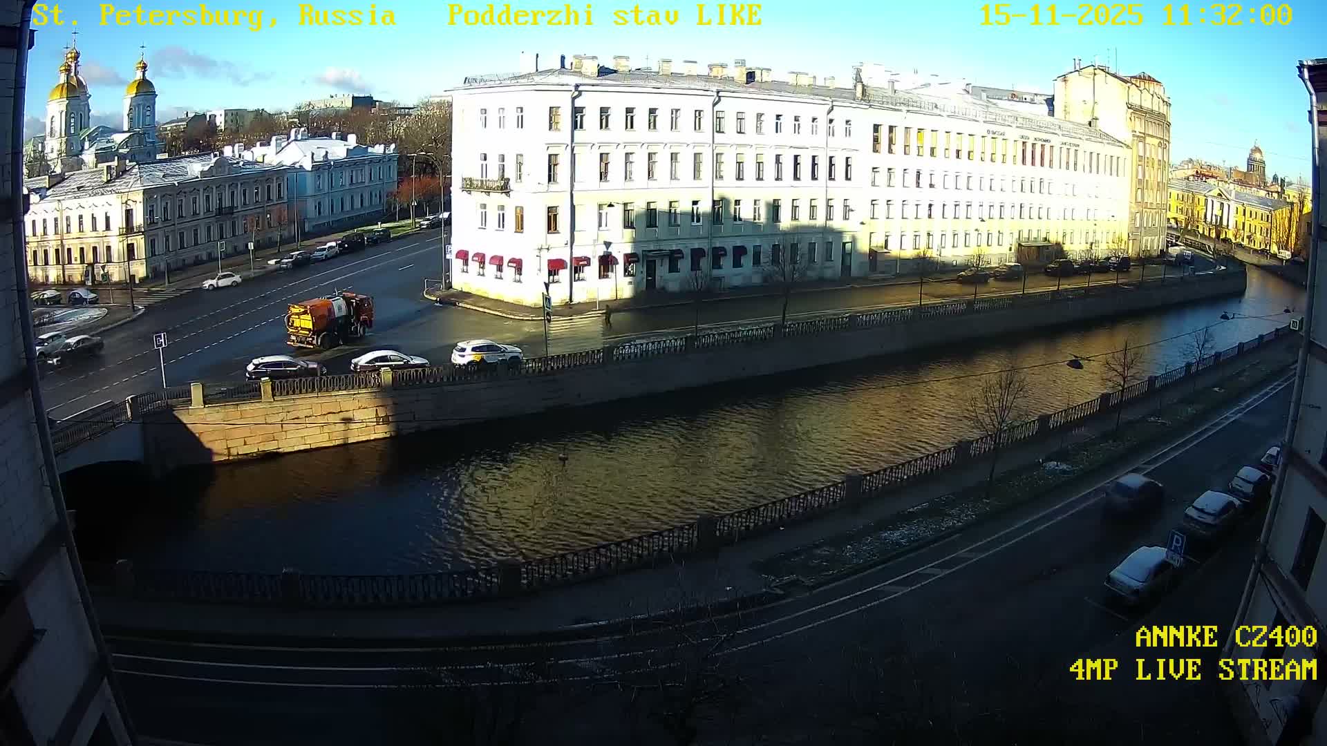A sunlit urban landscape unfolds with a canal winding between historic buildings and tree-lined embankments, traversed by roads carrying vehicles, all under a bright blue sky with scattered clouds.
