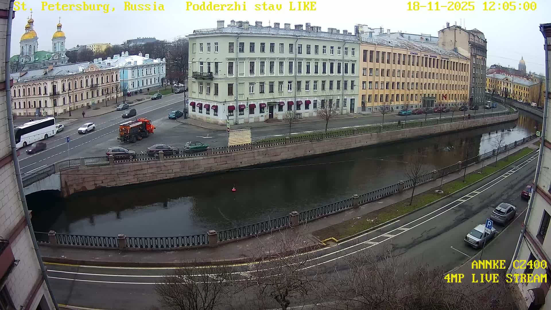 A sunlit urban landscape unfolds with a canal winding between historic buildings and tree-lined embankments, traversed by roads carrying vehicles, all under a bright blue sky with scattered clouds.