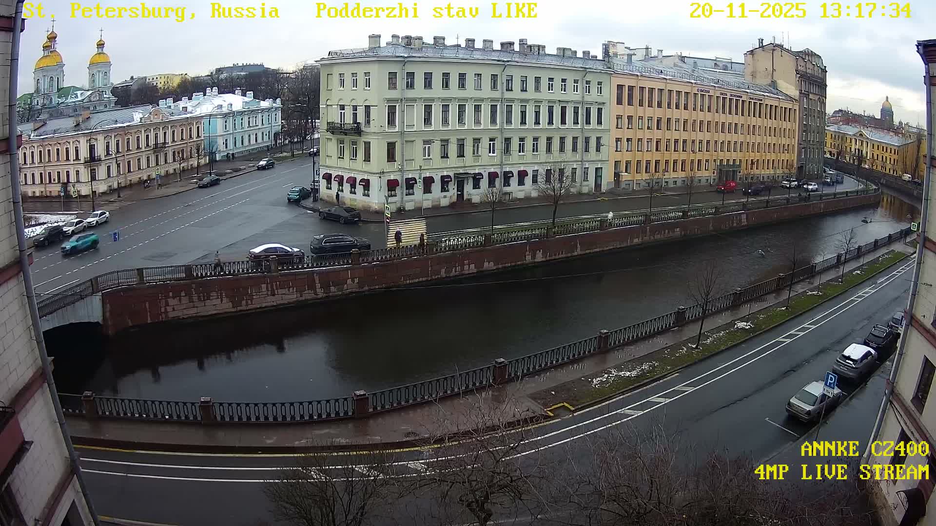 This wide-angle view captures a grey and wet urban scene with a canal flowing under a bridge, flanked by rows of classic European buildings and streets with parked and moving cars, all under an overcast sky with patches of snow on the ground.