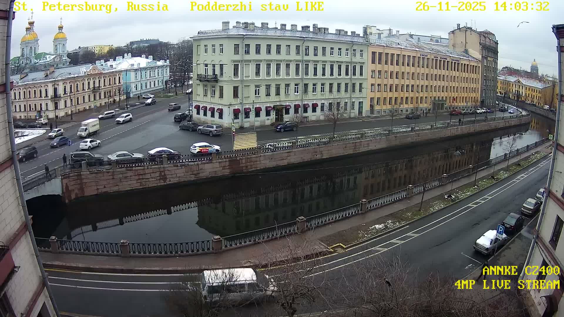 This wide-angle view captures a grey and wet urban scene with a canal flowing under a bridge, flanked by rows of classic European buildings and streets with parked and moving cars, all under an overcast sky with patches of snow on the ground.