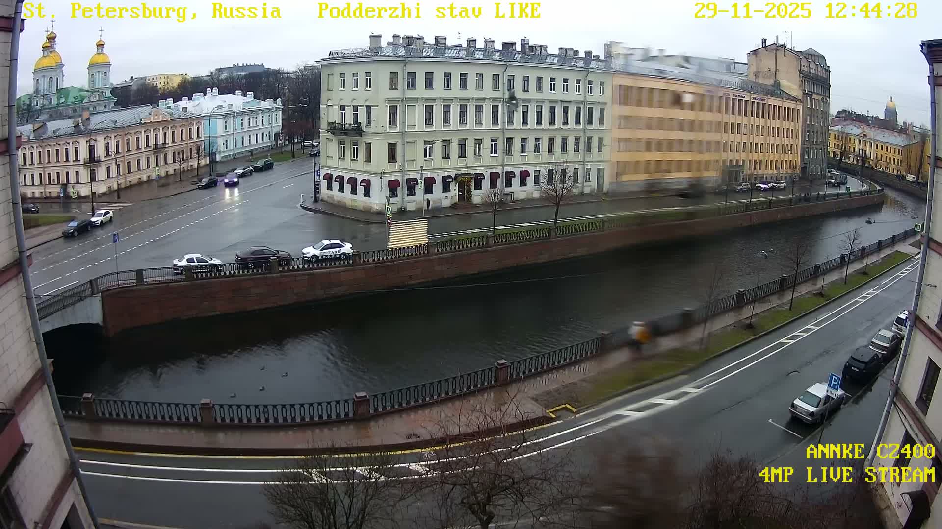 On a damp and overcast day, an aerial view shows a city canal flanked by wet roads busy with cars and lined with historical European-style buildings, including distant golden-domed structures, under a grey sky.