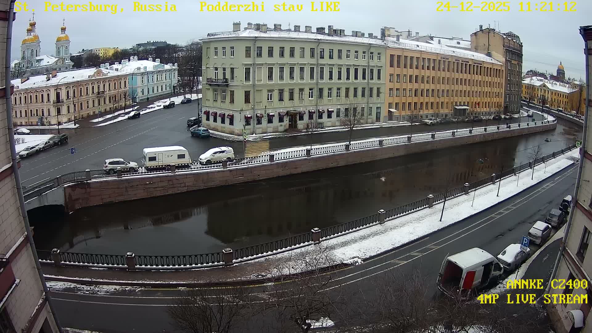 An overcast winter day reveals a snow-dusted city street running alongside a dark canal, bordered by multi-story buildings and occupied by various parked vehicles, including a white caravan on a bridge.