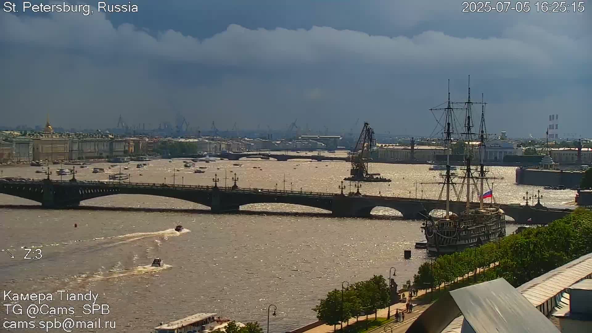 A large sailing ship is docked on a river next to a bridge, with a city skyline and partly cloudy skies in the background.