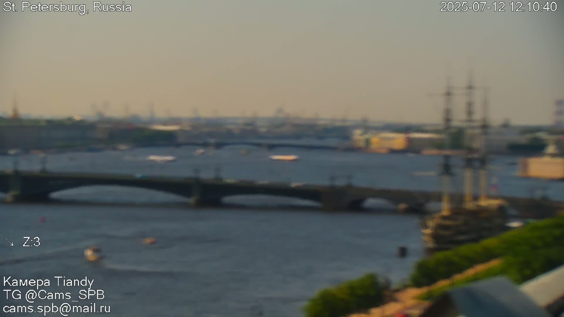 A large sailing ship is docked on a river next to a bridge, with a city skyline and partly cloudy skies in the background.
