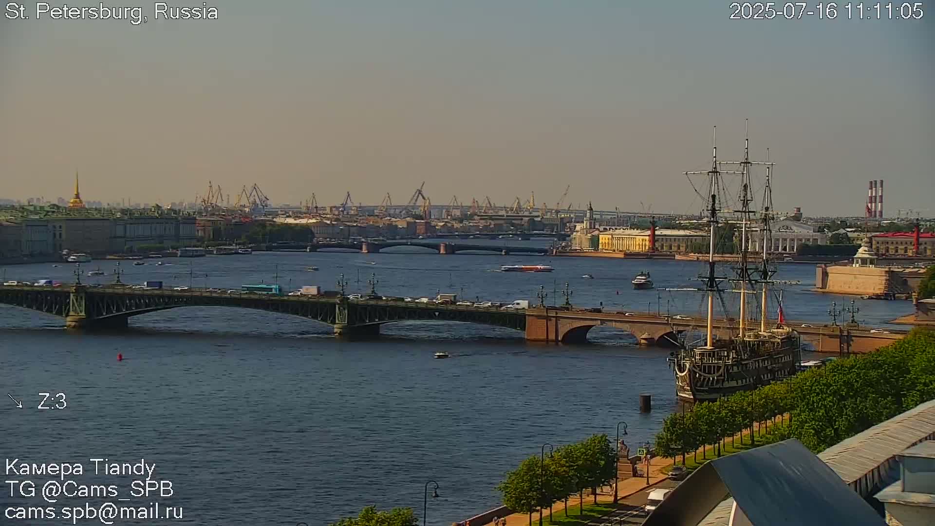 A large sailing ship is moored on a river, with bridges and city buildings visible in the background under a hazy sky.