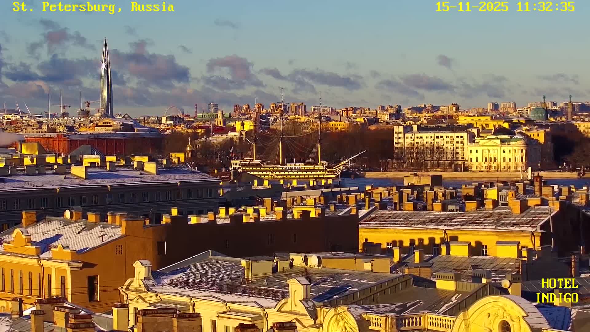 A sunny and partly cloudy winter cityscape reveals rooftops dusted with snow, golden-lit buildings, a tall modern skyscraper, and a historic sailing ship docked on a waterway.