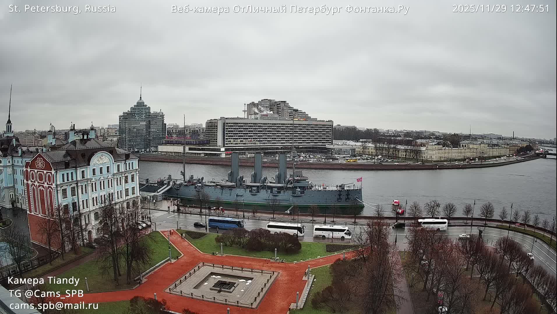 On an overcast and wet day, a historic gray cruiser is prominently docked on a wide river, with a bustling cityscape of various architectural styles and a park with bare trees and a wet road in the foreground.