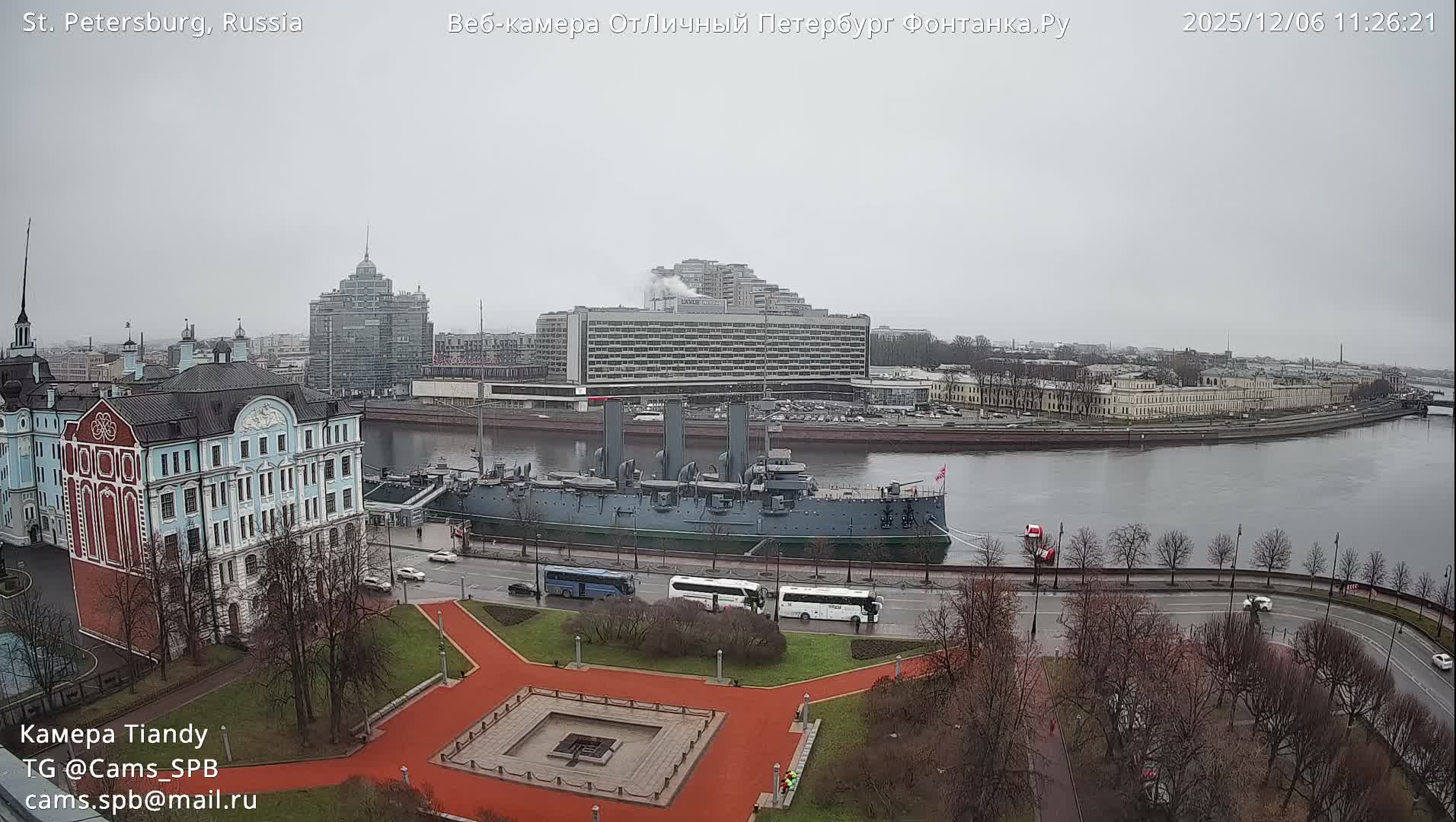 A historic grey cruiser is docked on a wide river under a heavily overcast and dreary sky, bordered by a park with bare trees, a busy road, and an urban skyline of diverse buildings.