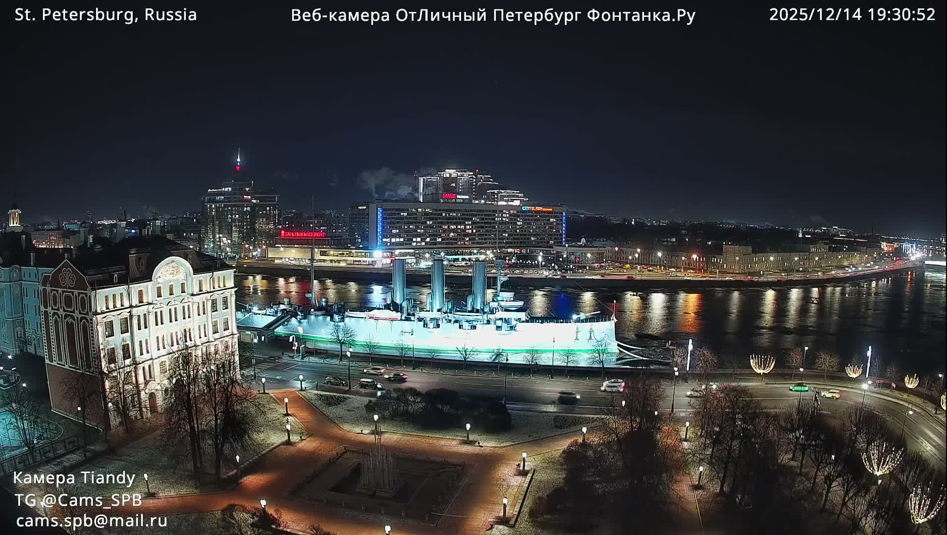 A historic grey cruiser is docked on a wide river under a heavily overcast and dreary sky, bordered by a park with bare trees, a busy road, and an urban skyline of diverse buildings.