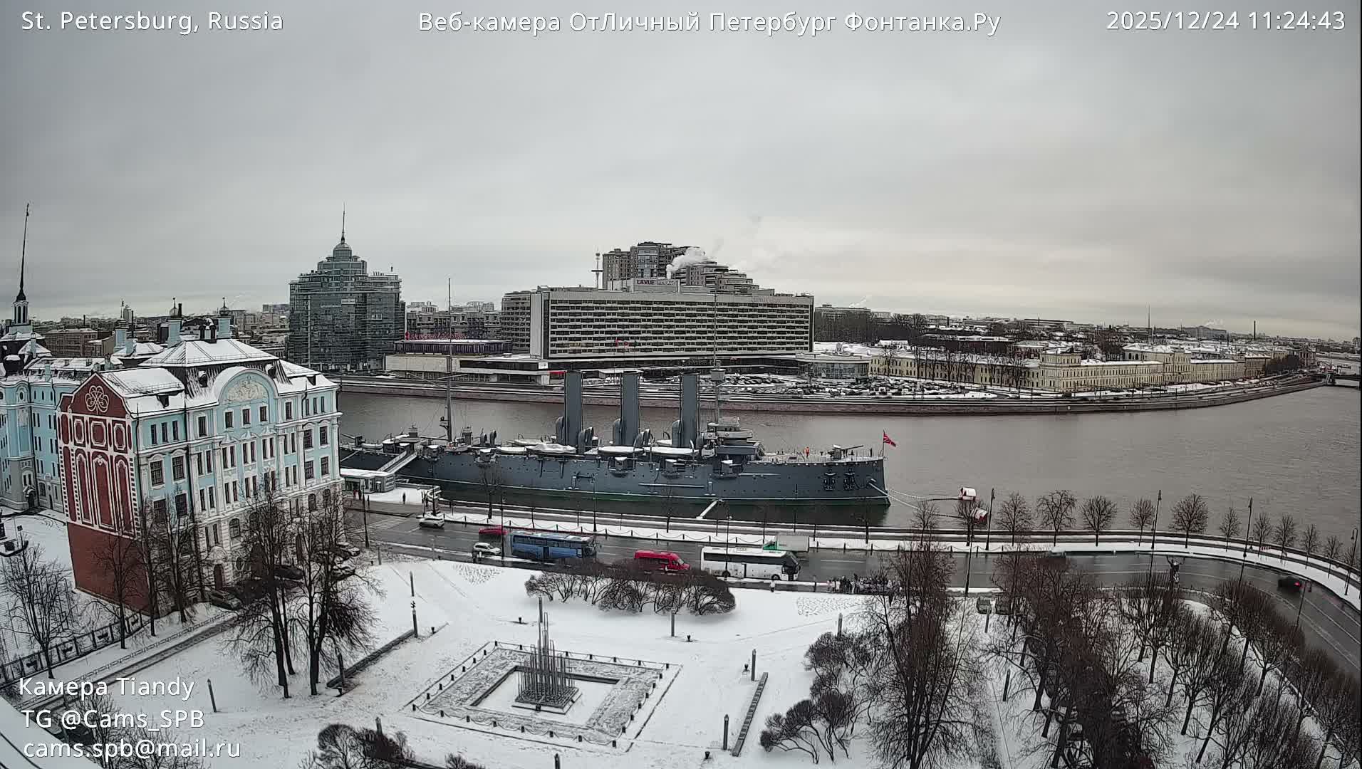 A large grey historic warship is docked on a river on an overcast winter day, with snow covering the ground, bare trees, and various city buildings lining the waterfront.