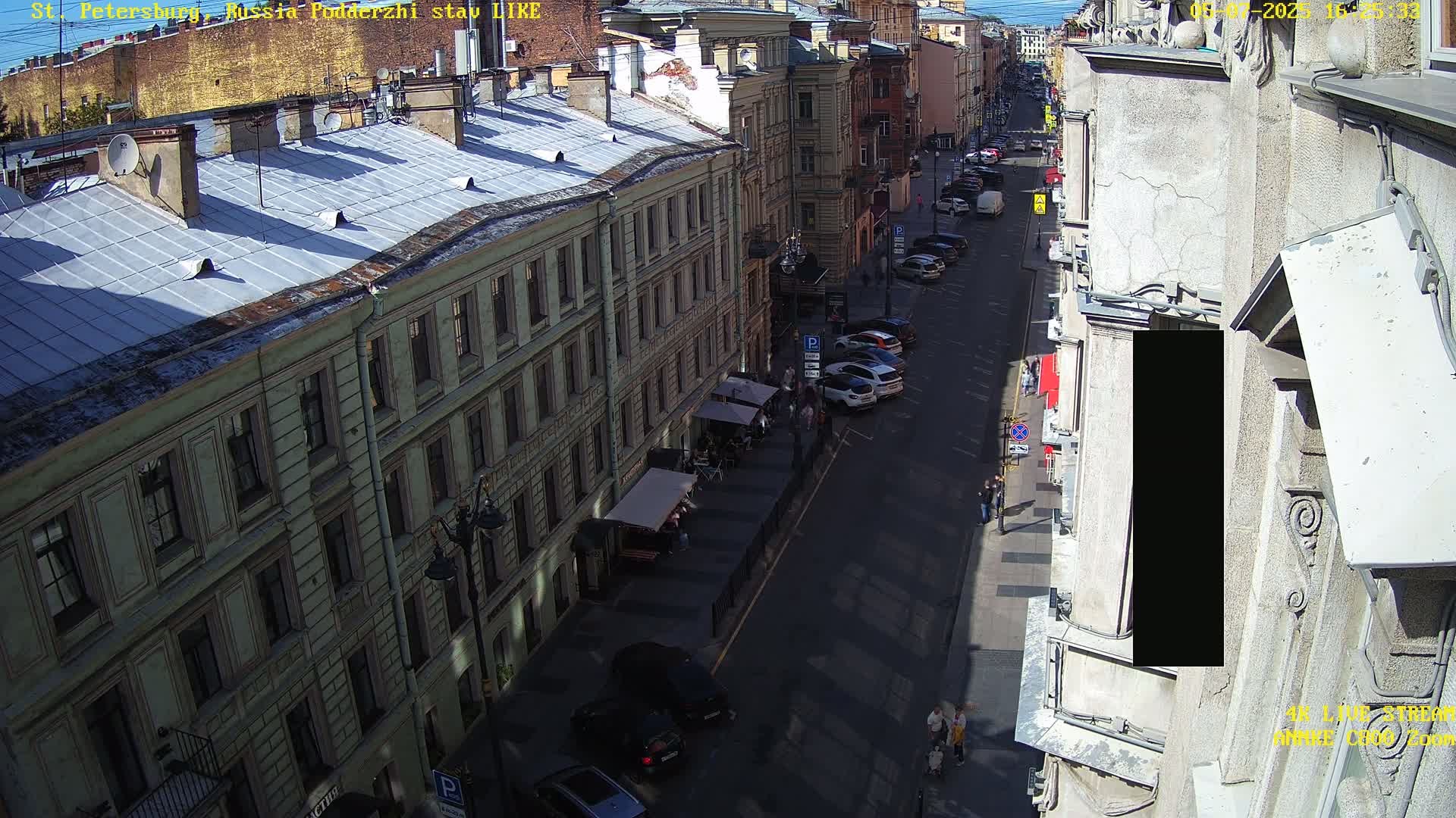 A sunny city street scene shows several parked cars lining a road between tall buildings with outdoor seating at a cafe.