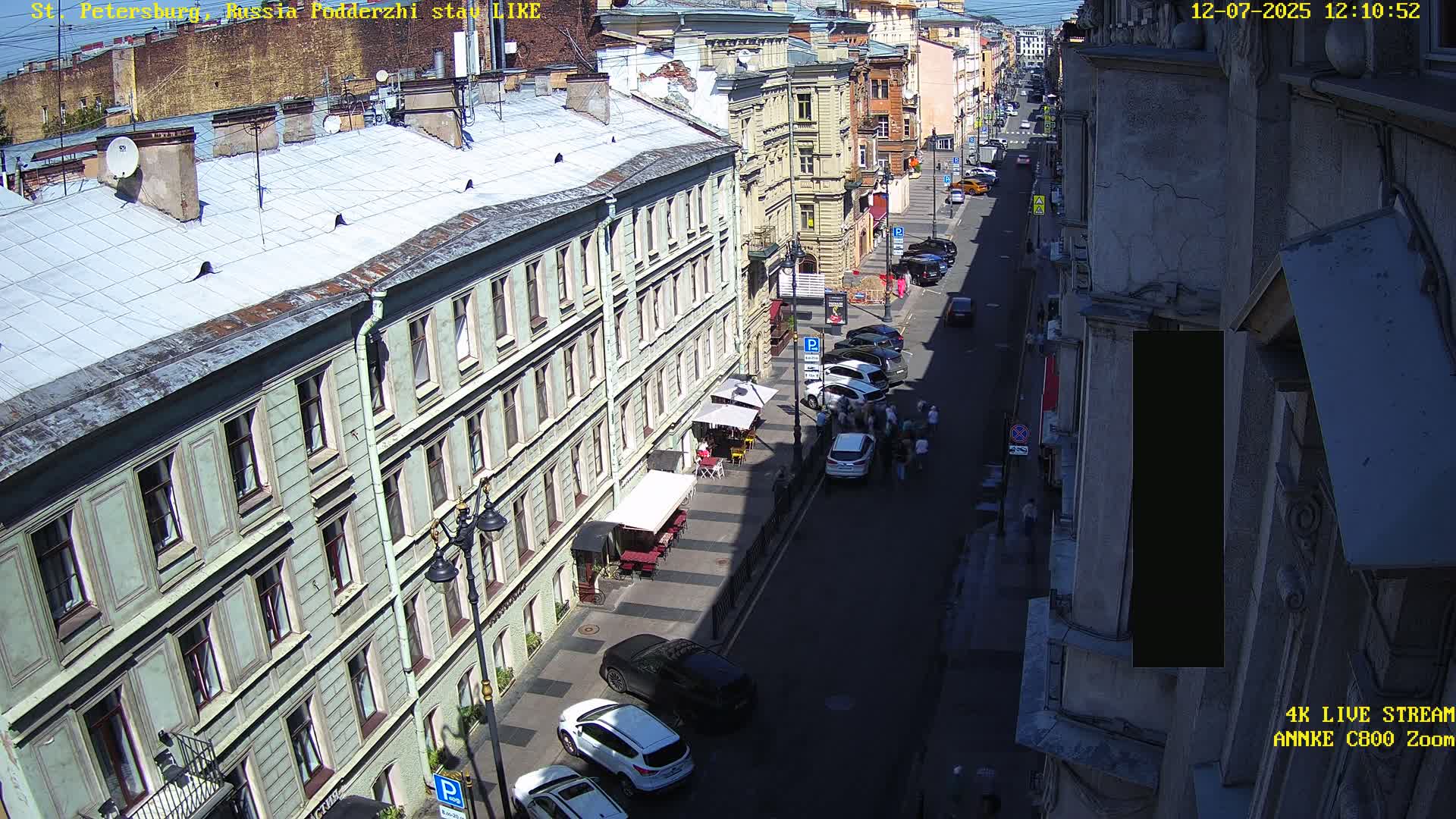 A sunny city street scene shows several parked cars lining a road between tall buildings with outdoor seating at a cafe.