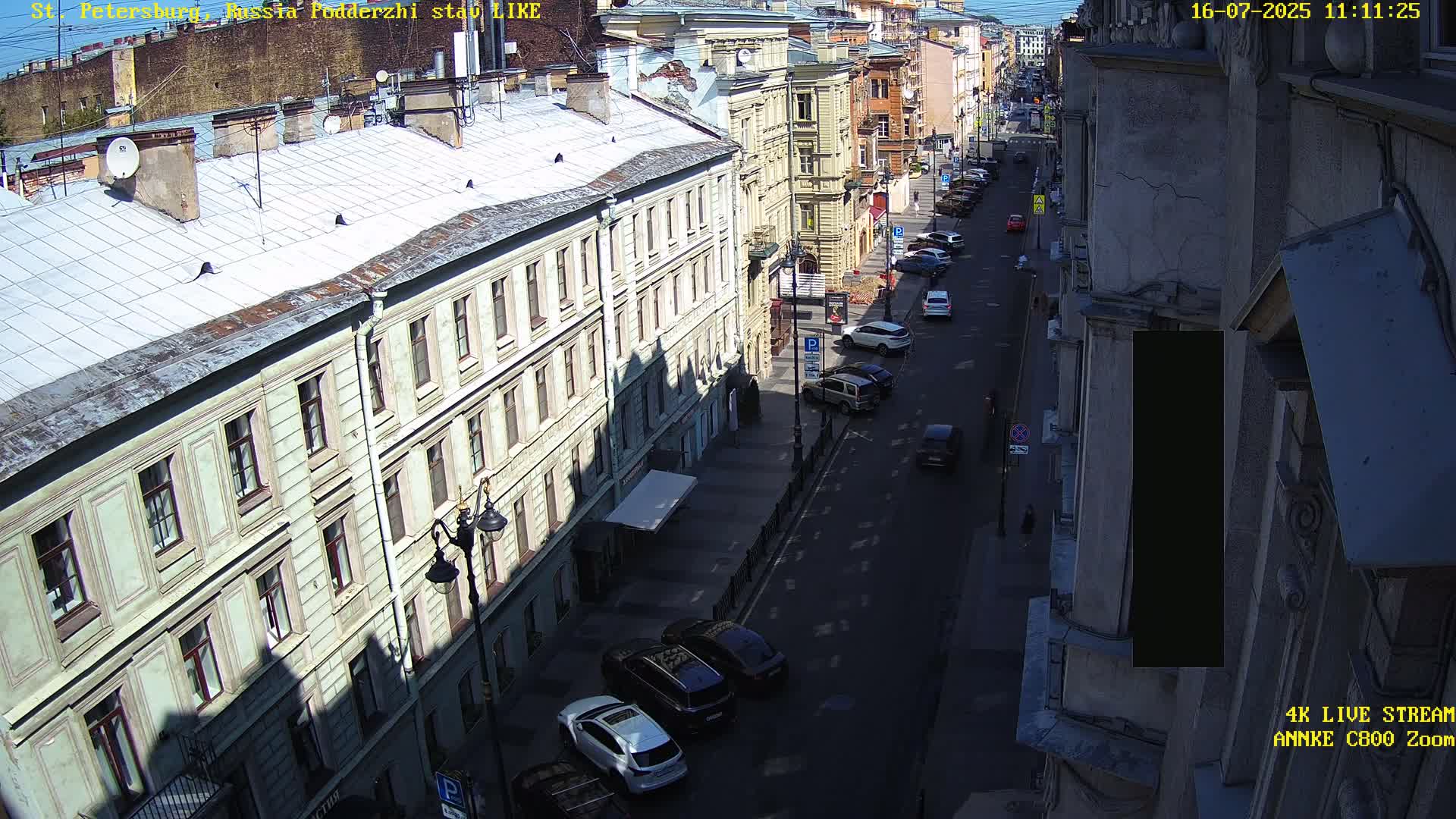 A sunny city street scene shows a row of multi-story buildings casting shadows on a street with parked and moving cars.