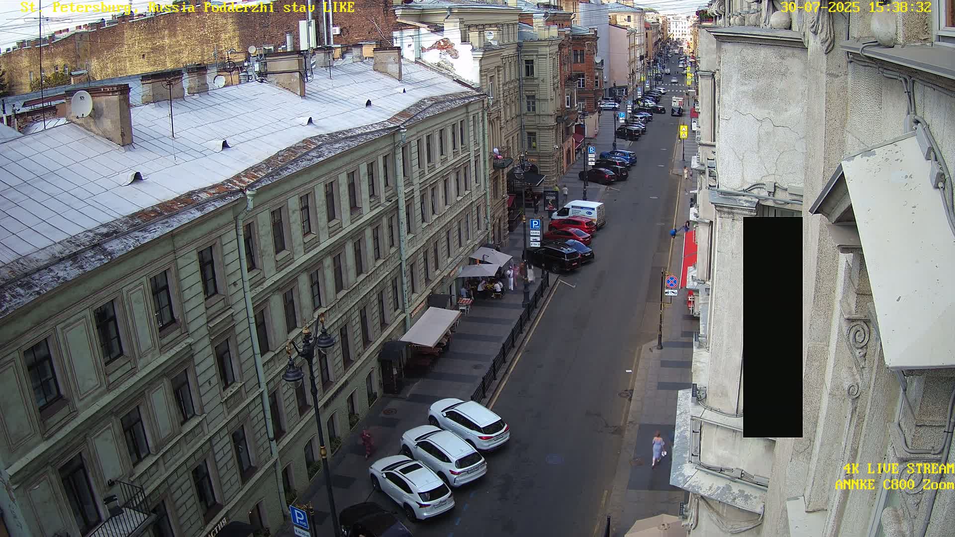 A sunny city street scene shows a row of multi-story buildings casting shadows on a street with parked and moving cars.