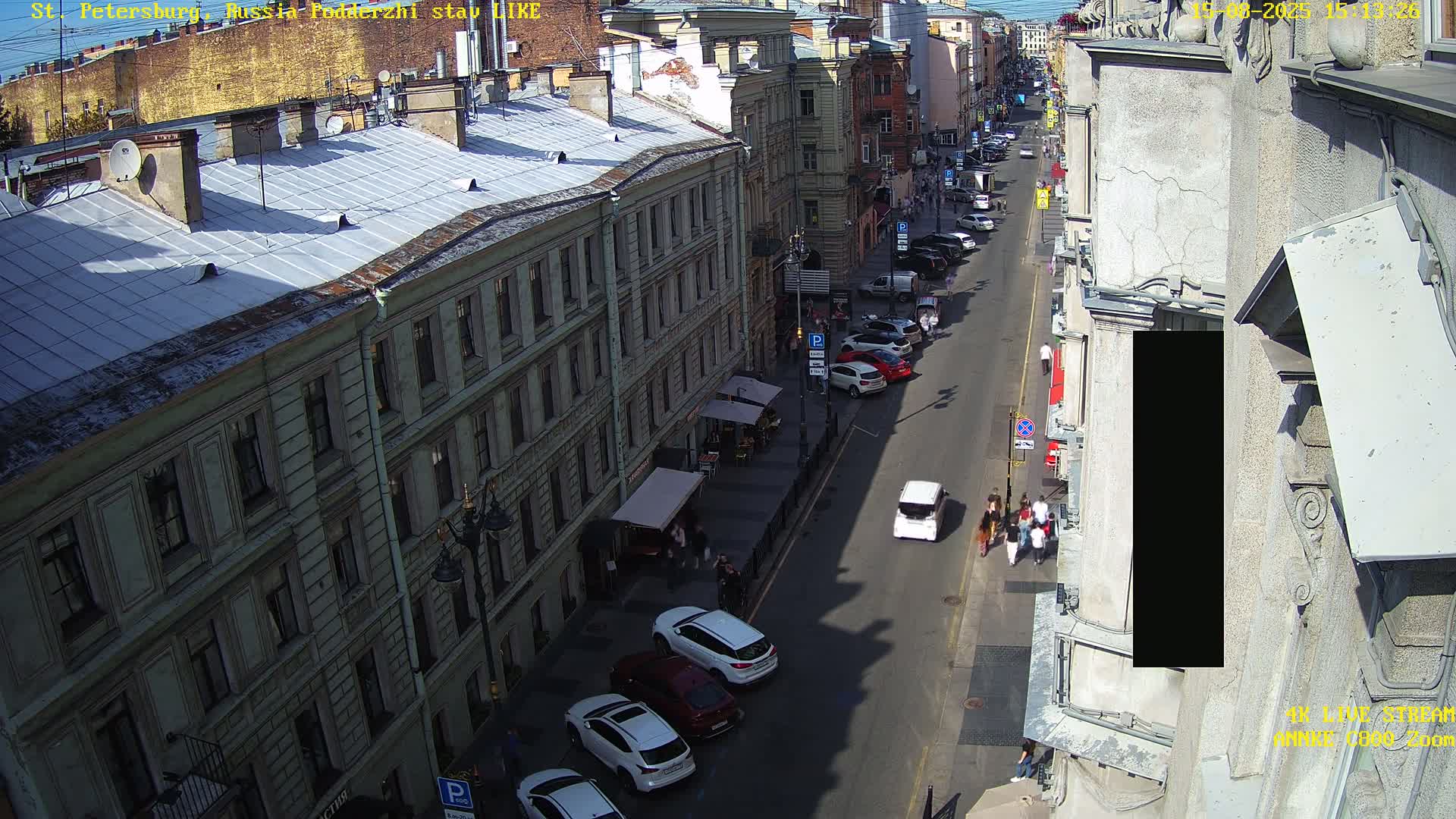 A sunny, high-angle view shows a city street with parked cars and pedestrians, flanked by multi-story buildings.