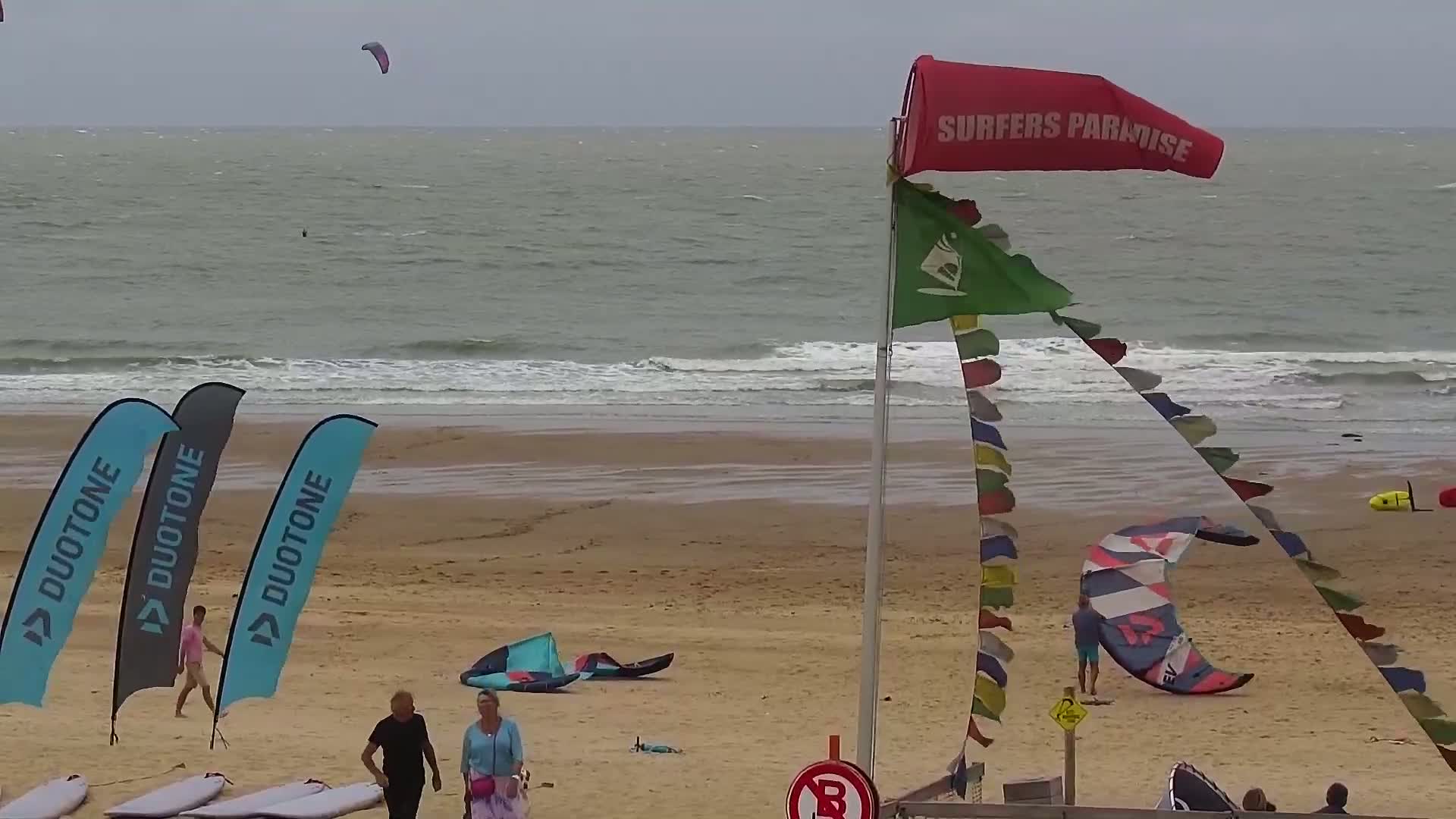 A sandy beach with several kitesurfers and their equipment, under an overcast sky with moderate waves.