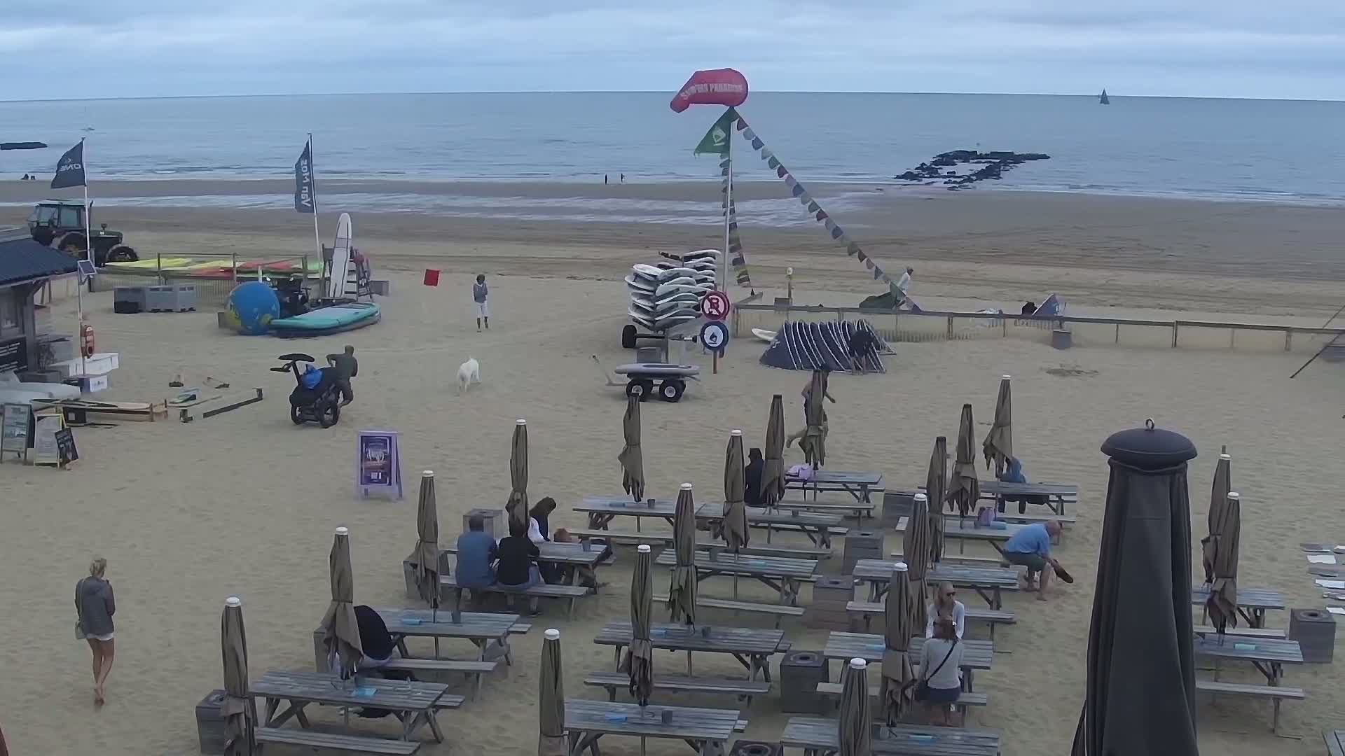 A sandy beach with several kitesurfers and their equipment, under an overcast sky with moderate waves.