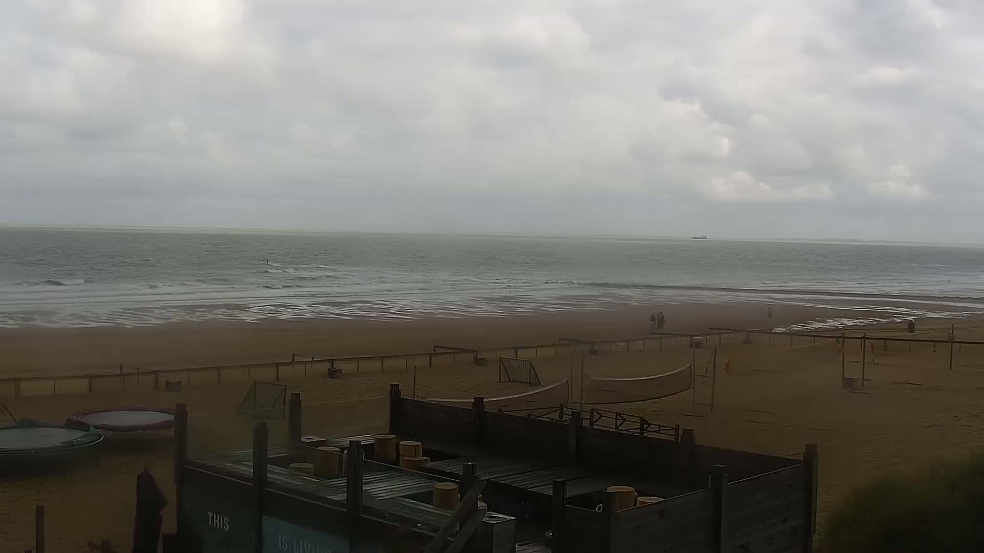 A mostly cloudy day at a beach shows a wide expanse of calm ocean water, a sandy beach with volleyball nets and a trampoline, and a few people walking.