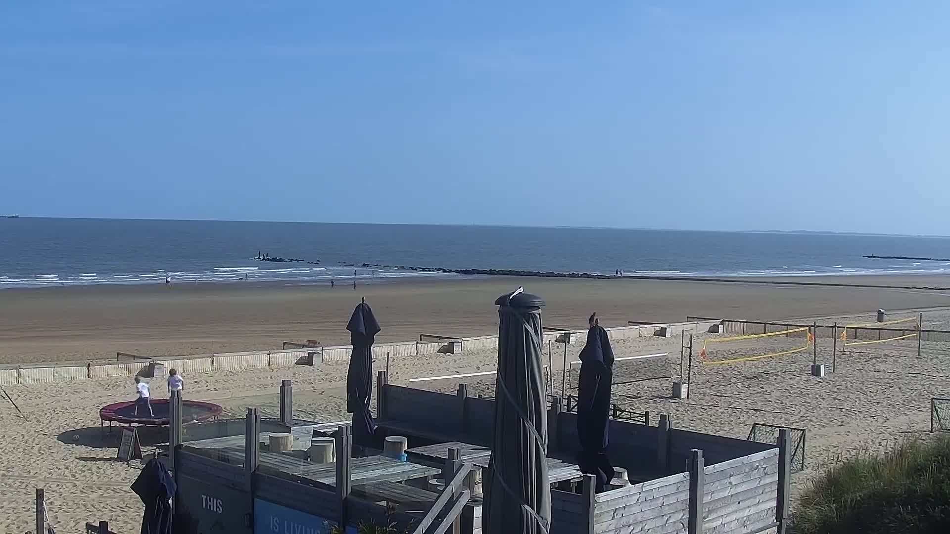 A sandy beach with calm ocean waves under a clear blue sky features beach volleyball courts, a trampoline with children playing, and a wooden structure with patio umbrellas.