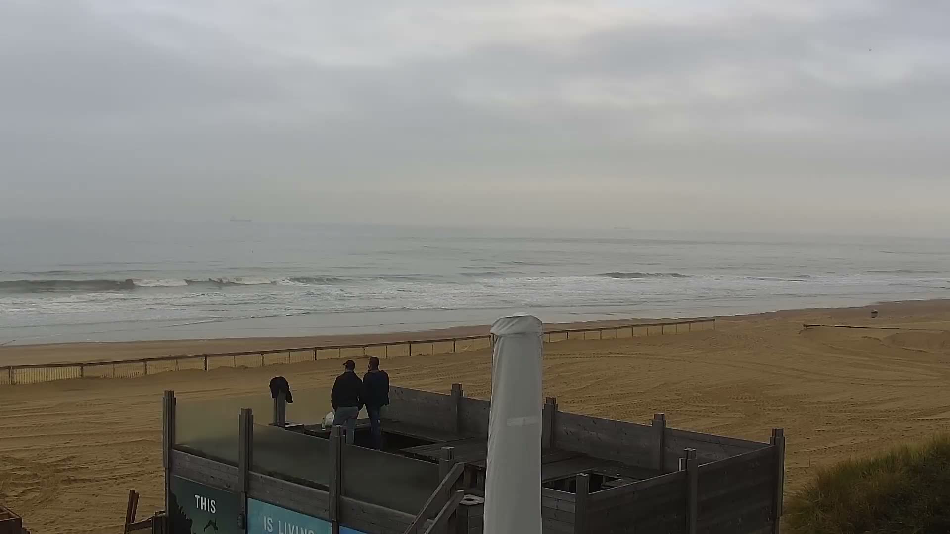 Two people stand on a wooden viewing platform overlooking a wide, sandy beach and a grey, wavy ocean under an overcast sky.